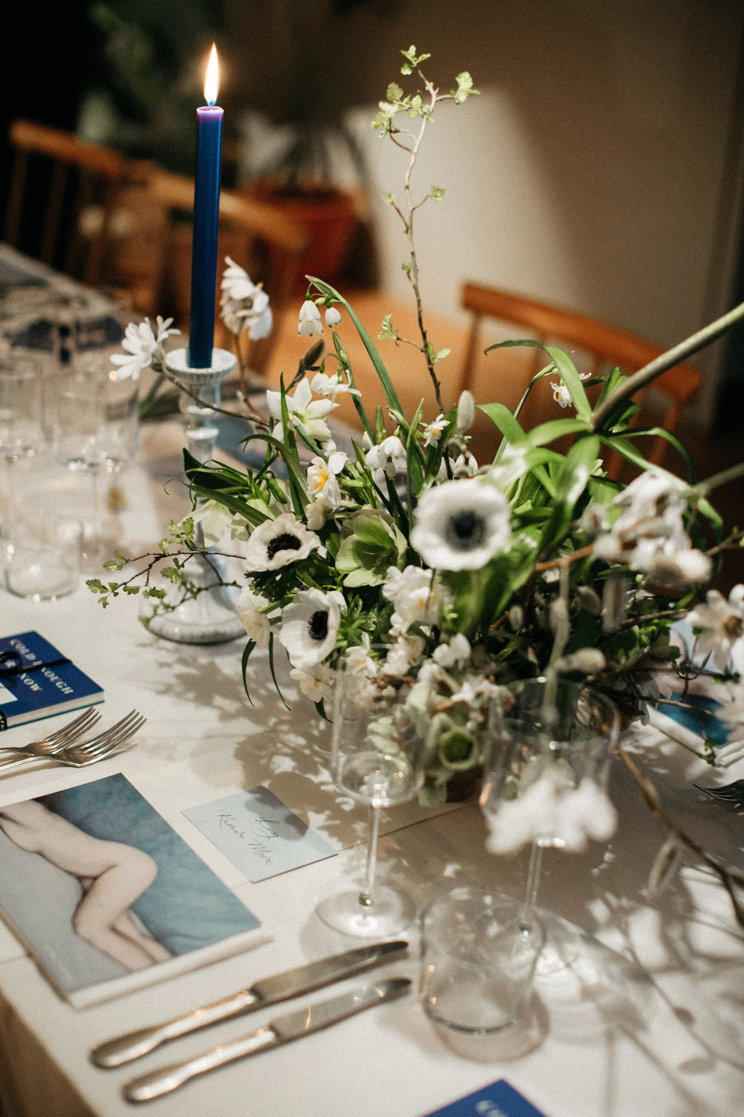 Elegant table setting with blue candle, floral centerpiece, glassware, and place cards on white tablecloth.
