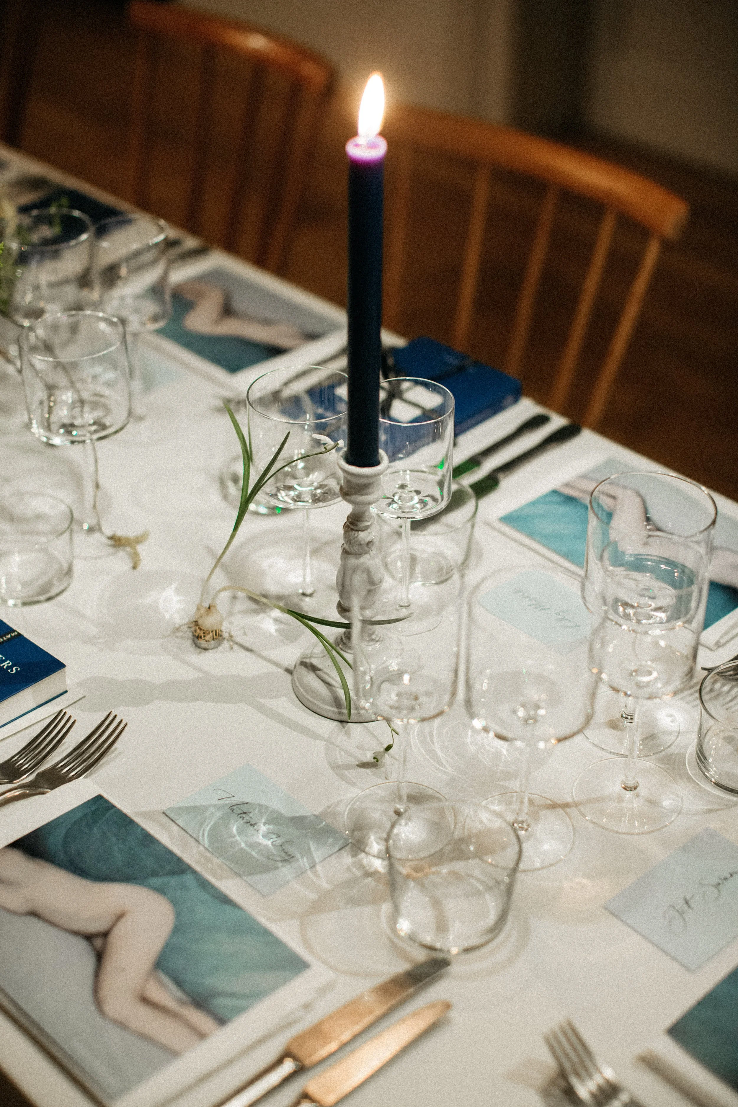 Elegant dining table setting with a tall lit blue candle, glassware, silverware, books, and artistic photography on a white tablecloth.