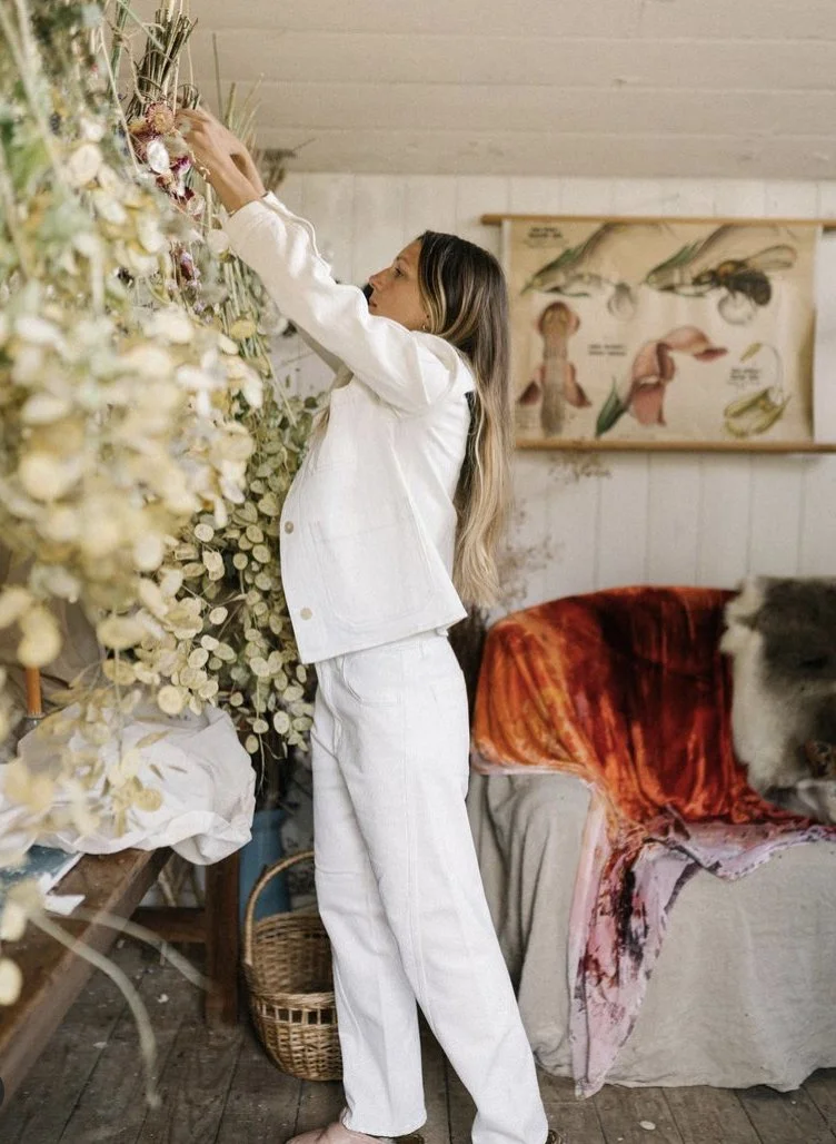 Milli Proust arranging dried flowers indoors, dressed in white, with a botany poster and cozy decor in the background.