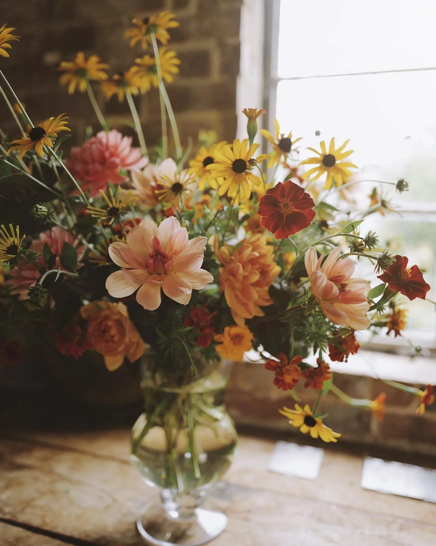 A glass vase with a colorful bouquet of flowers including pink dahlias, yellow daisies, and red blossoms, placed on a wooden table near a window.