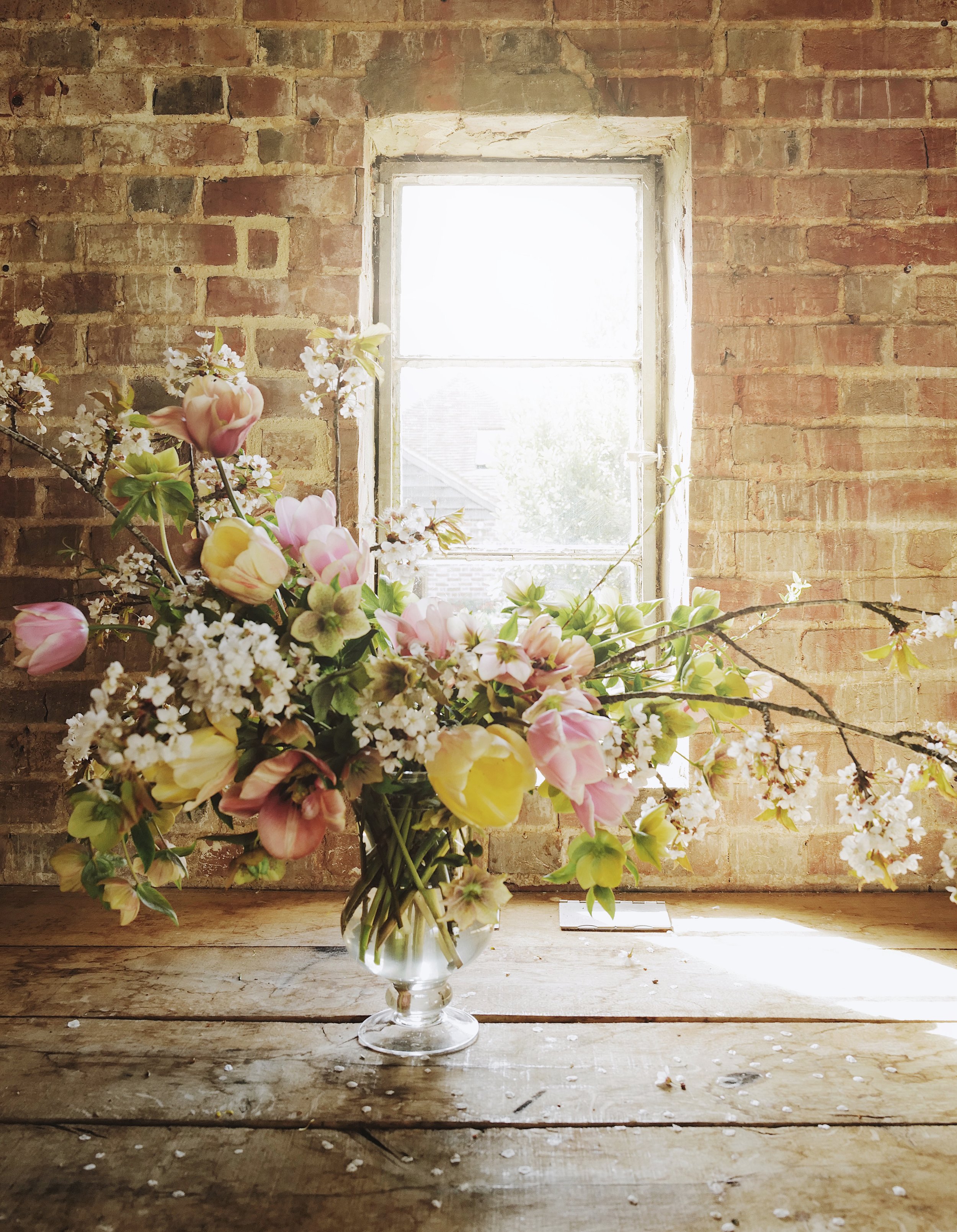 Flower arrangement with tulips and cherry blossoms in glass vase on wooden table near brick wall and window.