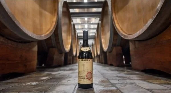 A bottle of red wine standing upright between large wooden wine barrels in a cellar.