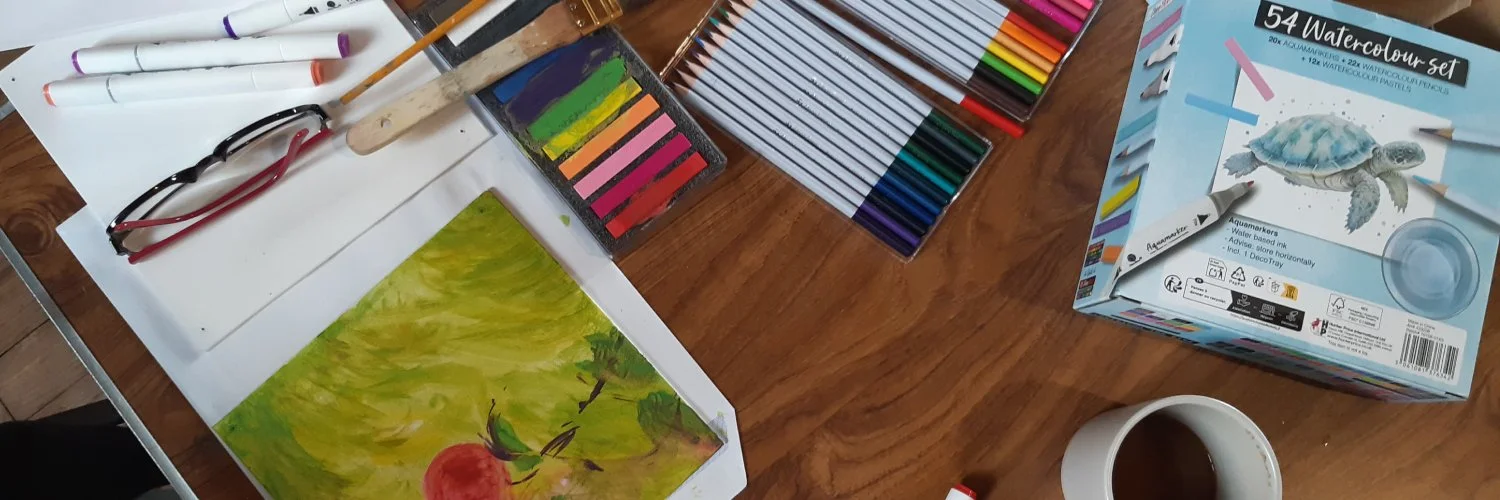 Partially painted artwork surrounded by art materials, brushes, pens, watercolours and a cup of tea.