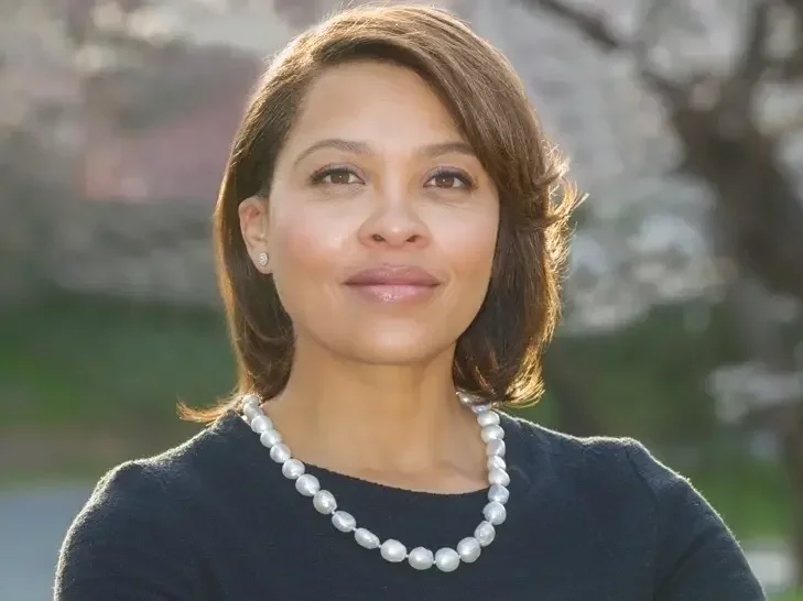 A woman with medium skin tone, short brown hair, and pearl earrings, wearing a black top and a pearl necklace, standing outdoors.