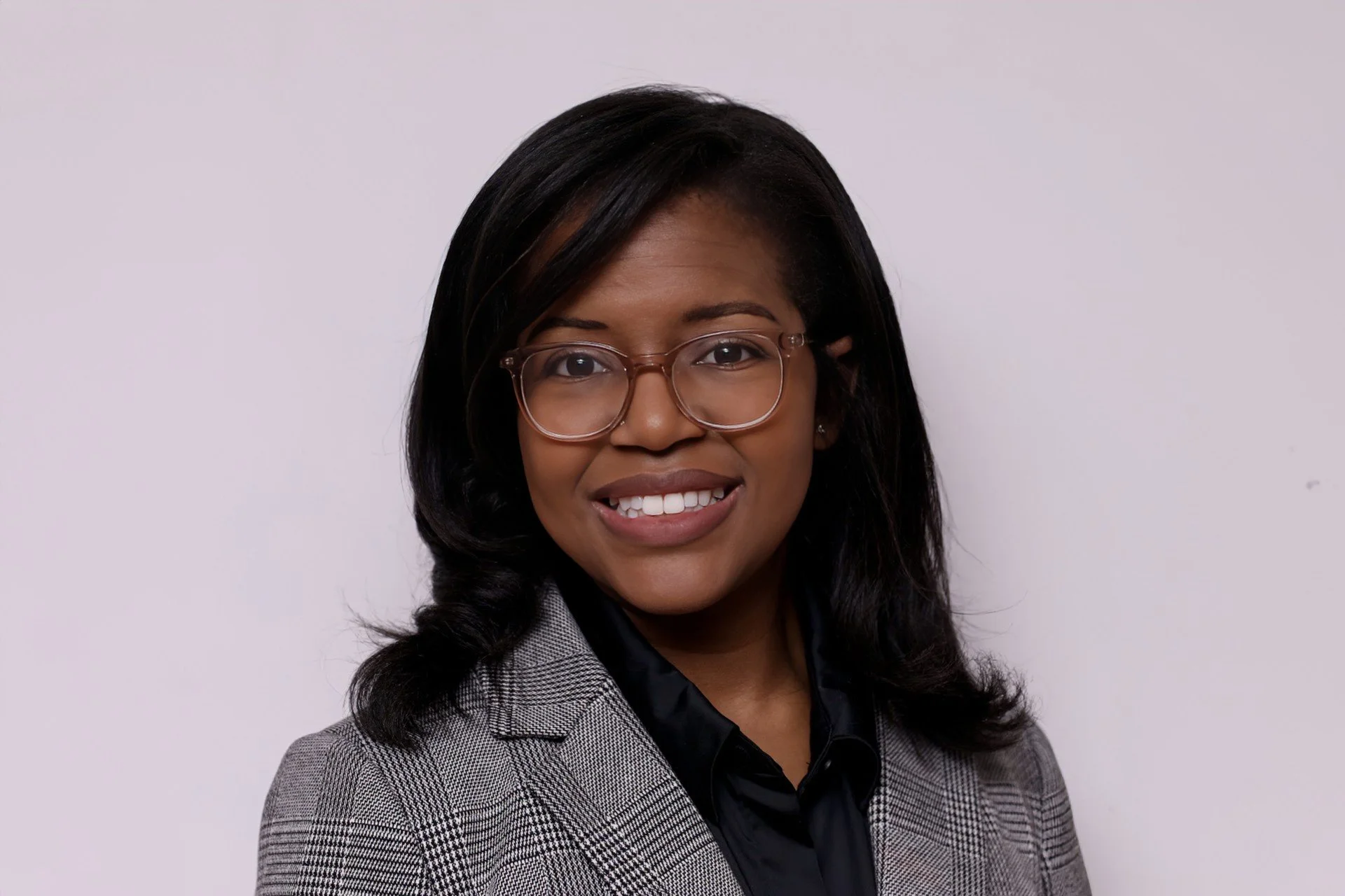 A woman with long hair, glasses, and a big smile, wearing a grey blazer and a black shirt against a plain light-colored wall.