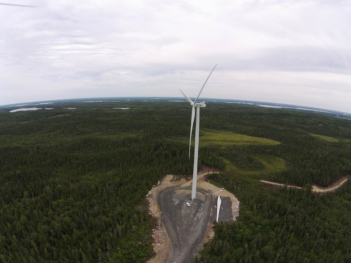 A wind turbine under construction in a forested area with roads and a cloudy sky.