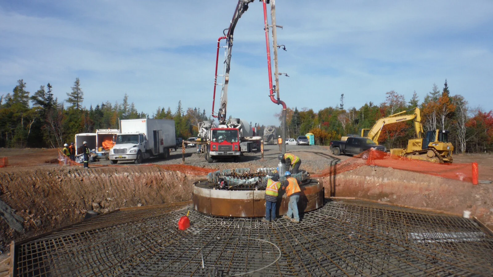 Construction workers on site pouring concrete into a large, circular rebar framework for a structure, with machinery and trucks in the background.