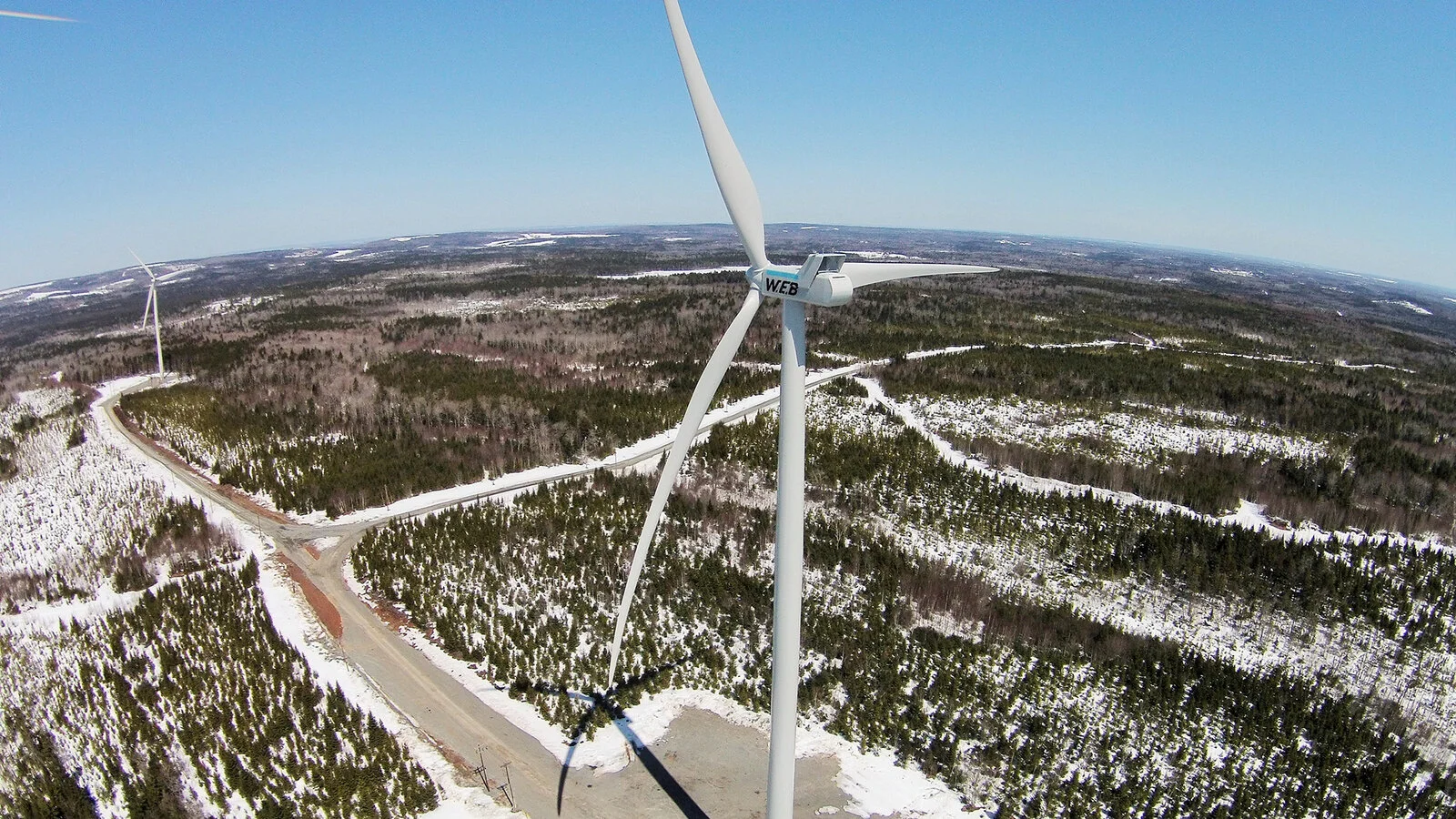 An aerial view of a wind farm with snow-covered fields and roads, showing several wind turbines, one in the foreground with three blades, in a winter landscape.