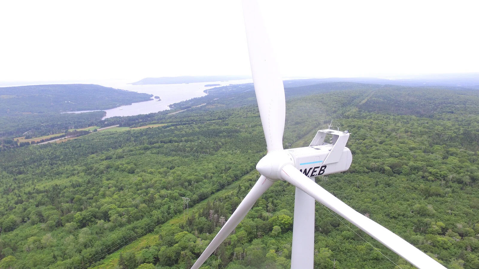 A white wind turbine with three blades spinning above a green forested landscape with a river and distant hills.