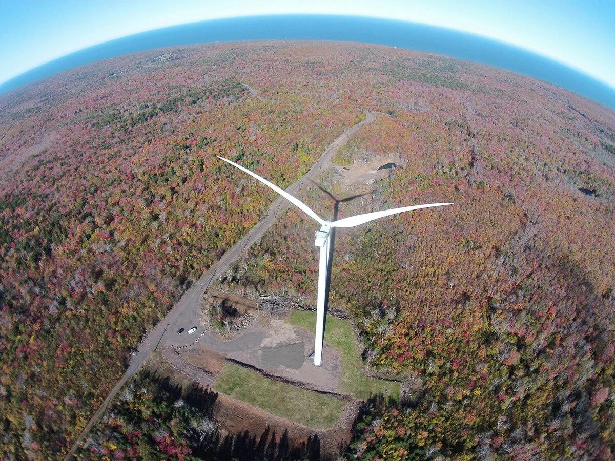 A large wind turbine situated in a forest with trees showing fall colors, under a clear blue sky. The image appears to be taken from a drone or aerial device, with the landscape curving around the horizon.