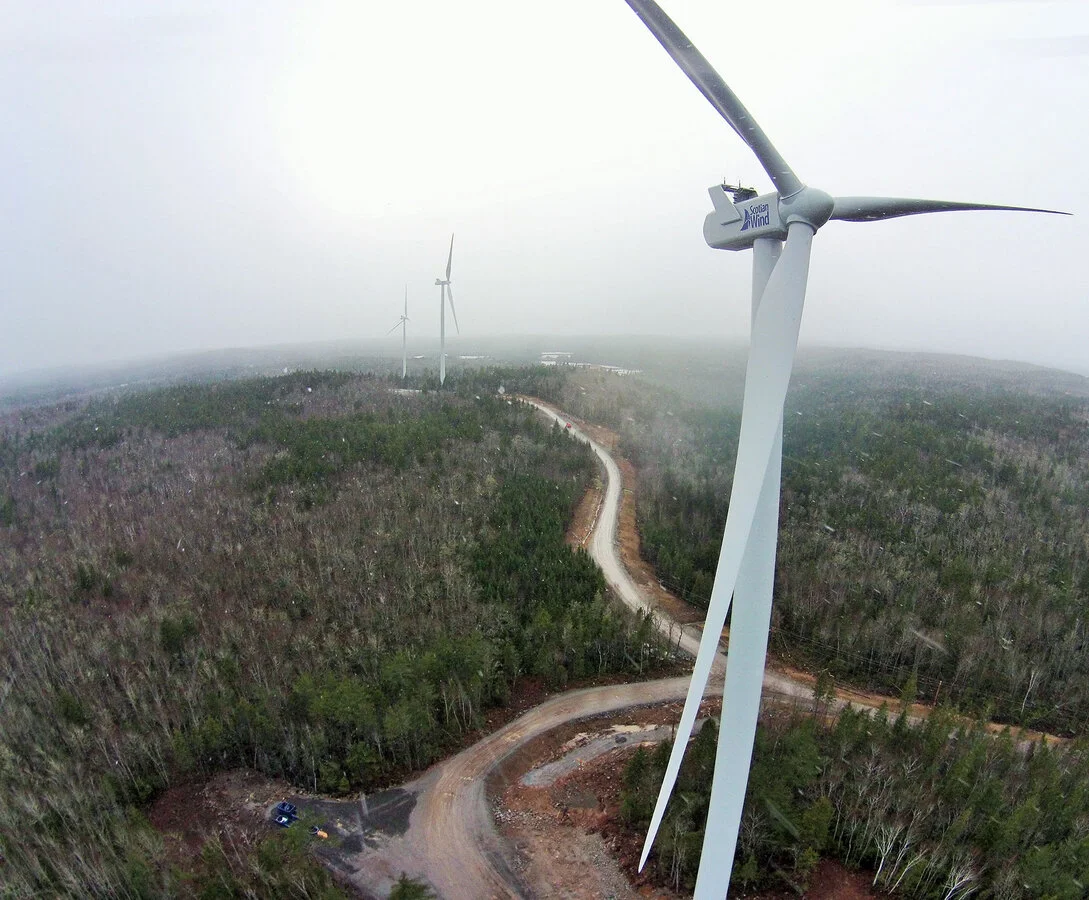 Aerial view of a wind farm with multiple wind turbines in a forested area and a winding dirt road. The closest turbine shows the blade broken and leaning.