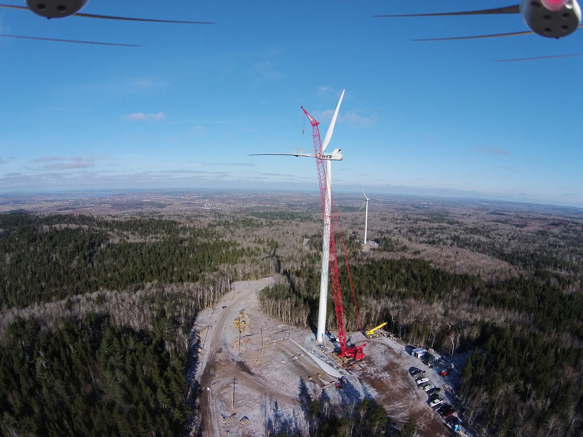 A wind turbine under construction with a crane next to it, in a forested area on a clear day.