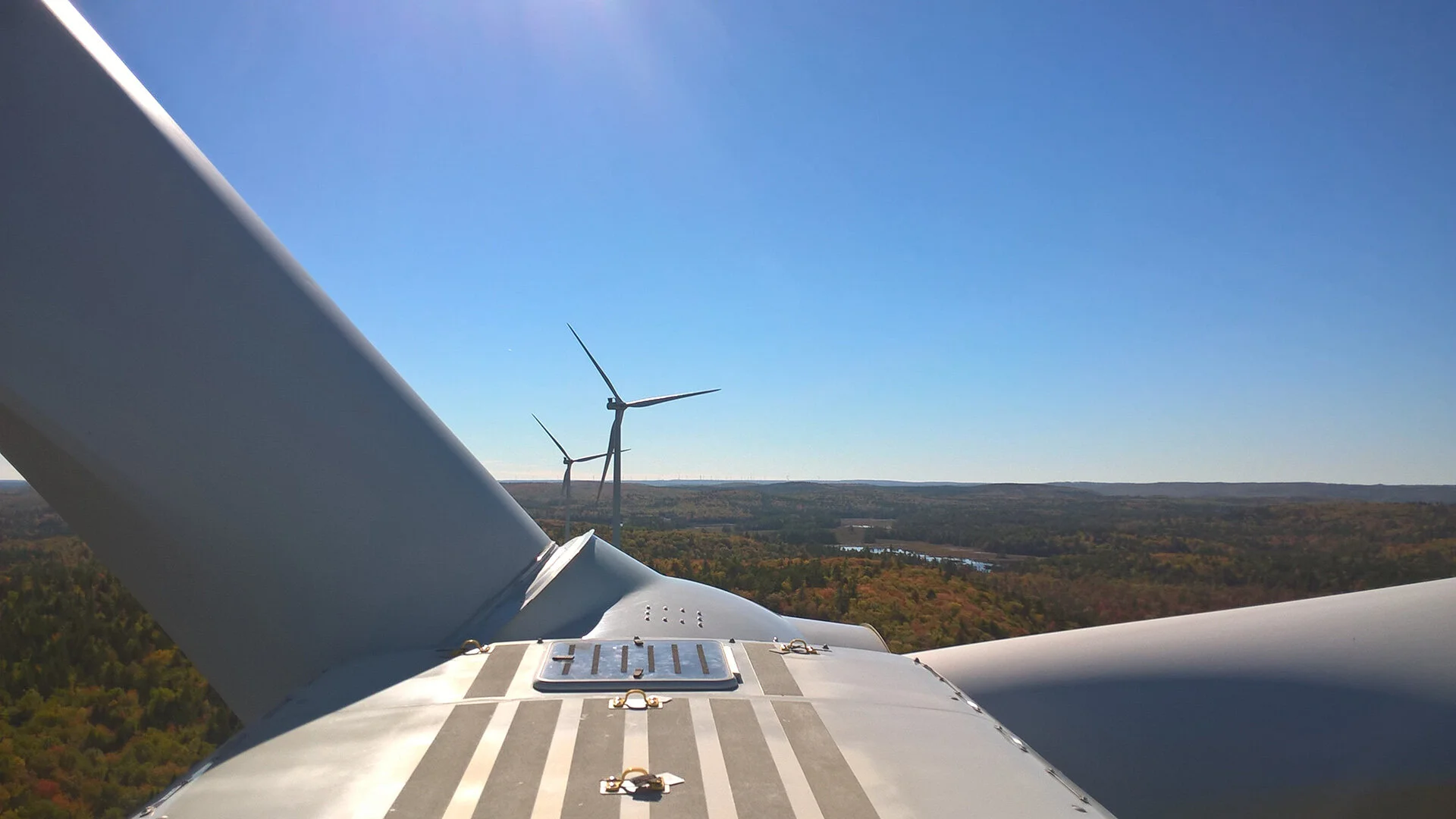 View from above of wind turbines on a hilltop, with a clear blue sky and a landscape of trees and a river in the distance.