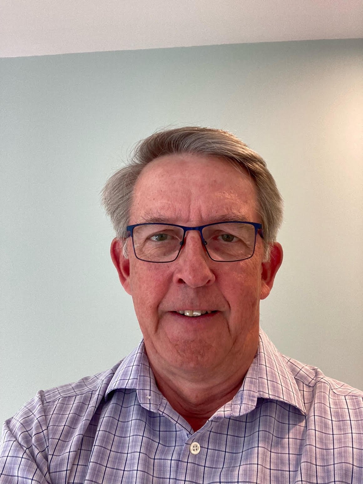 A middle-aged man with gray hair and glasses smiling, wearing a checkered shirt, against a plain light-colored wall.