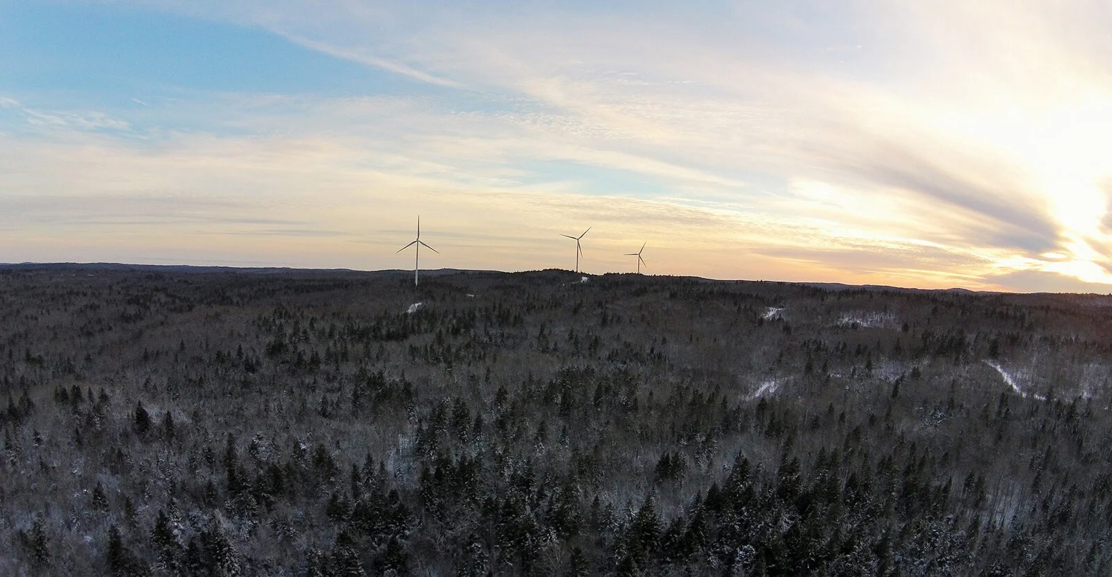 A landscape of a snow-covered forest with wind turbines on a hill at sunset.