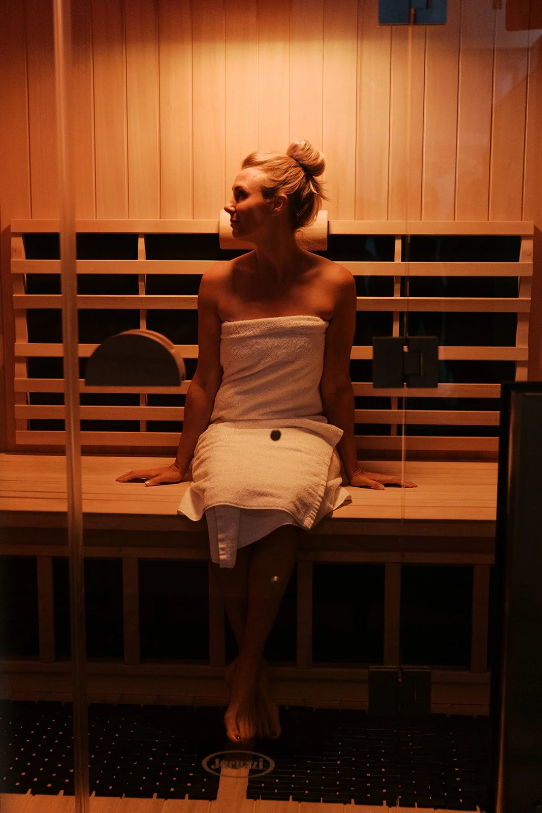 A woman wrapped in a towel sitting in a sauna, looking to her left, with wooden walls and benches around her.