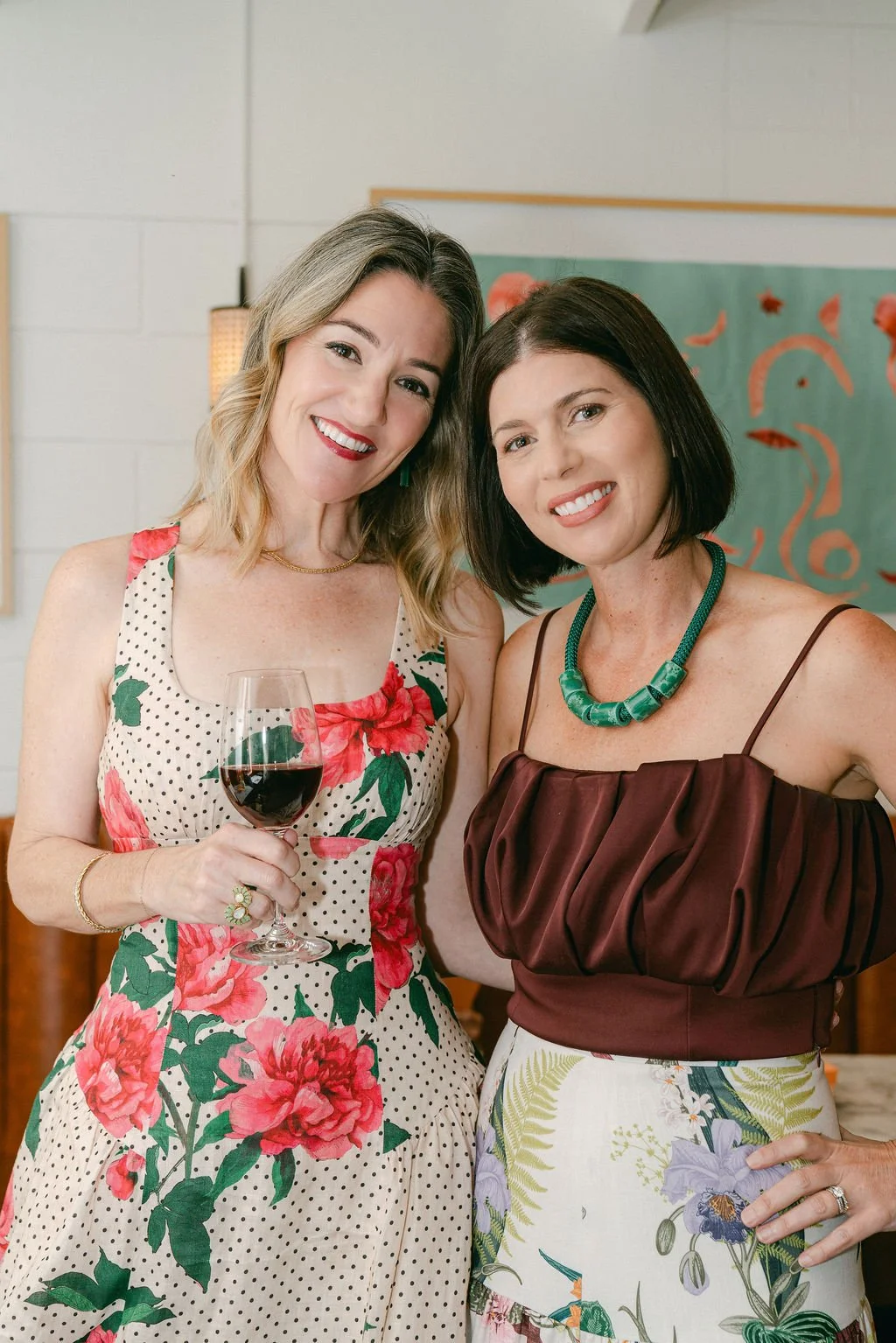 Two women smiling and posing for a photo indoors, one holding a glass of red wine. The woman on the left has blonde hair and is wearing a sleeveless floral dress, while the woman on the right has dark hair and is wearing a burgundy top with a green necklace and a floral skirt.