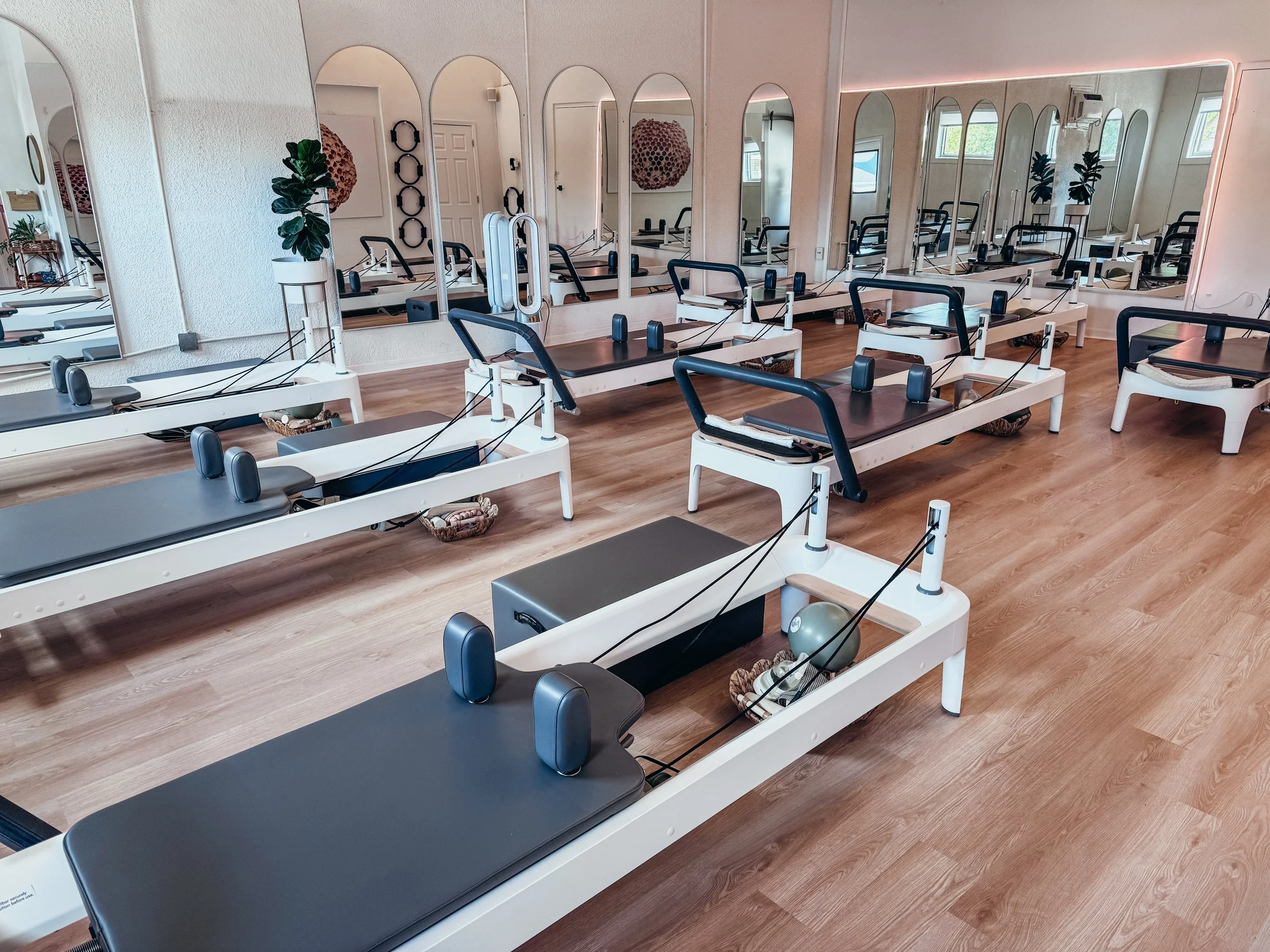 Pilates reformer machines in a bright, minimalistic exercise studio with mirrors on the walls and several windows.