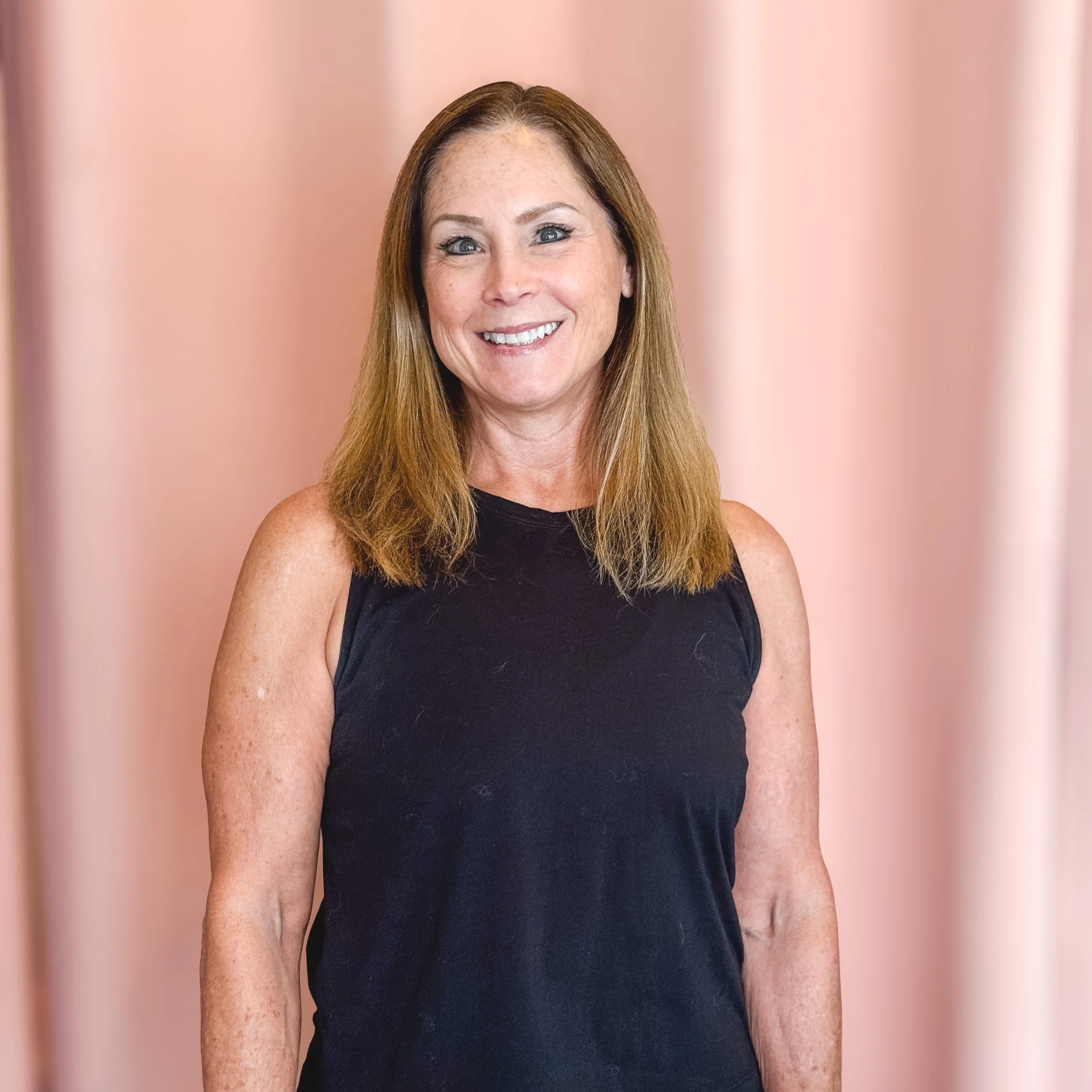 A woman with long red hair, fair skin, and freckles, smiling while wearing a black sleeveless top, standing in front of a pink background.