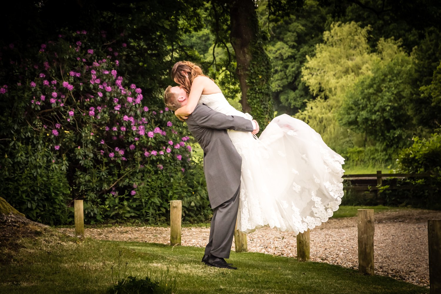 A man, wearing a gray suit, lifting a woman in a white wedding dress in a garden with green trees and pink bushes.