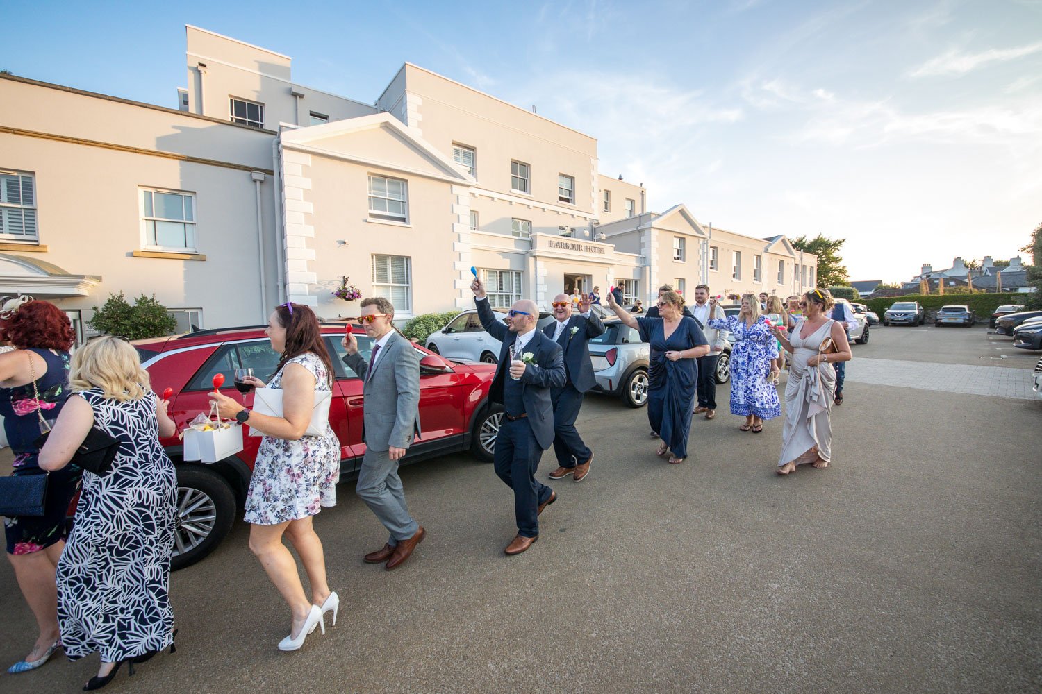 Wedding guests in formal attire walking out of a hotel with drinks and glasses of wine, on a sunny day.