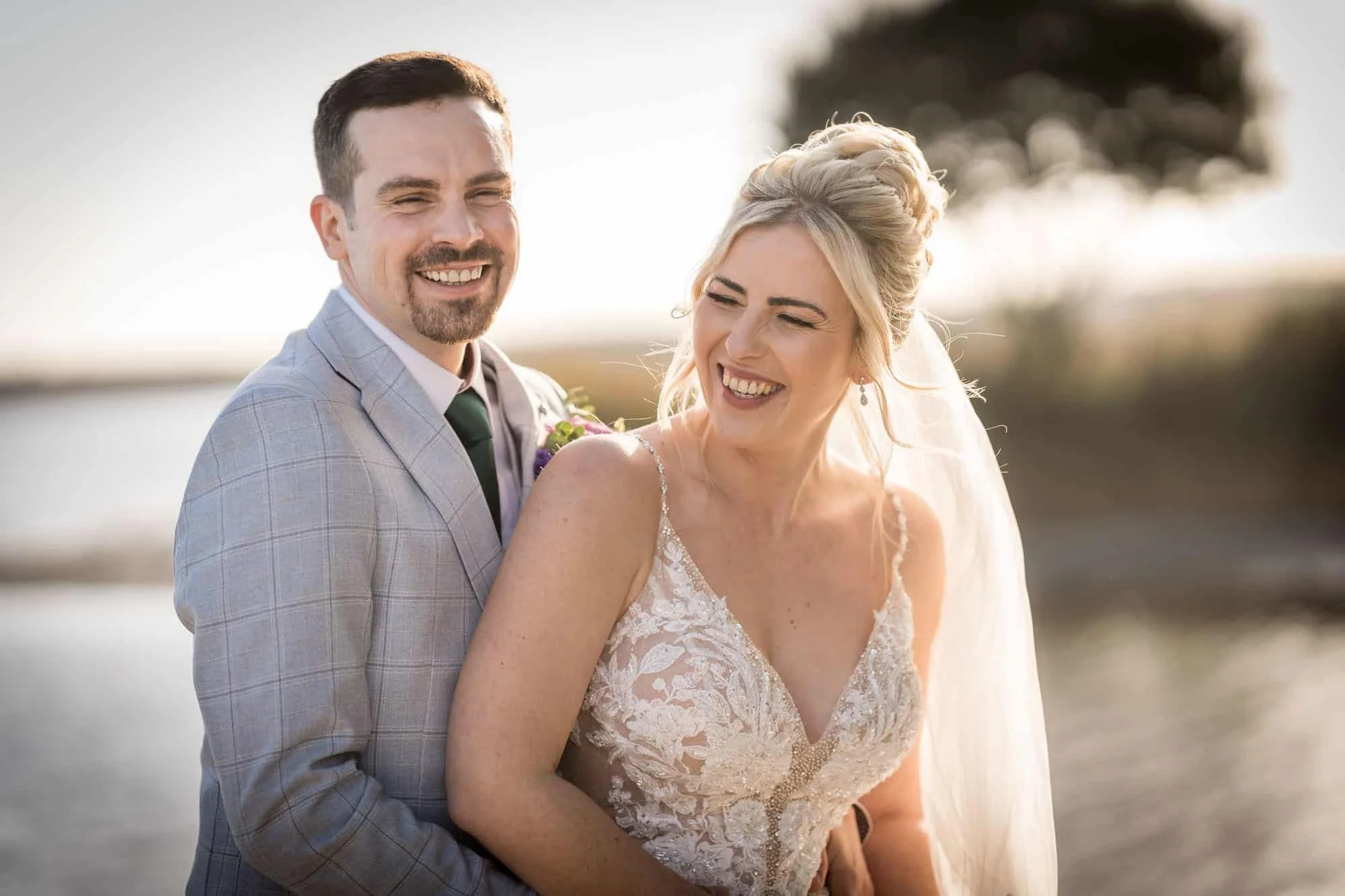 A happy bride and groom smiling outdoors during sunset at a wedding, with water and trees in the background.