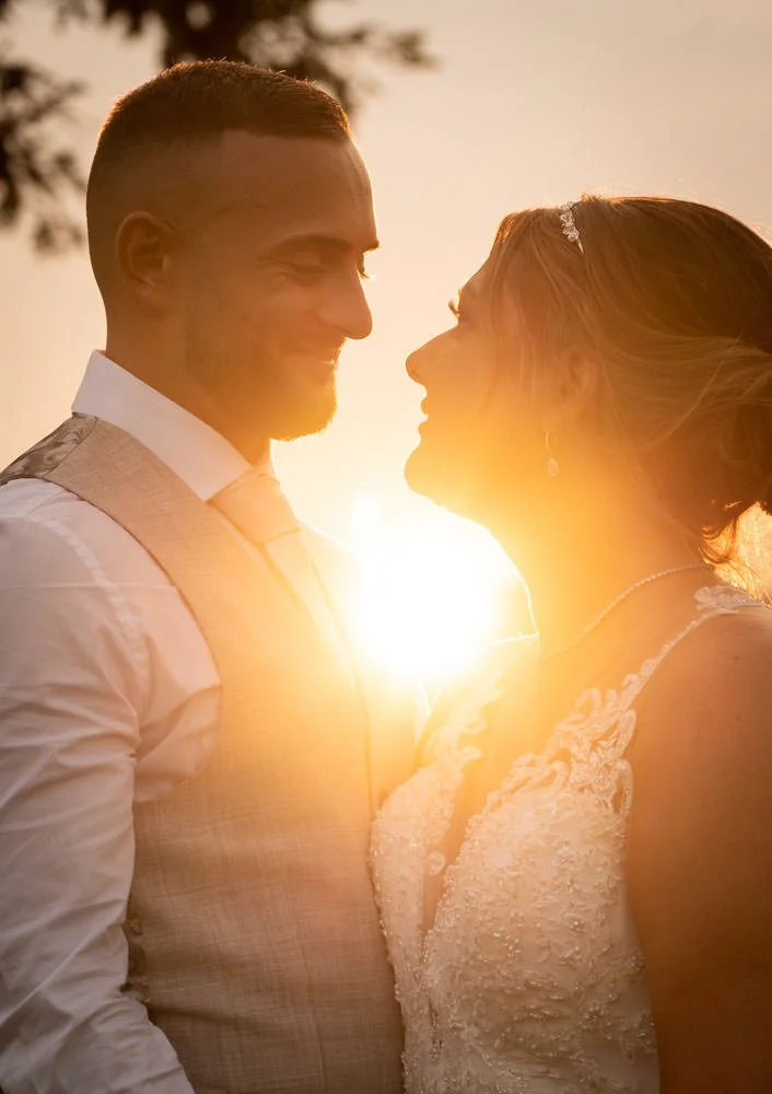 A groom and bride stand close together, smiling at each other during sunset, with the sun behind them creating a warm glow.