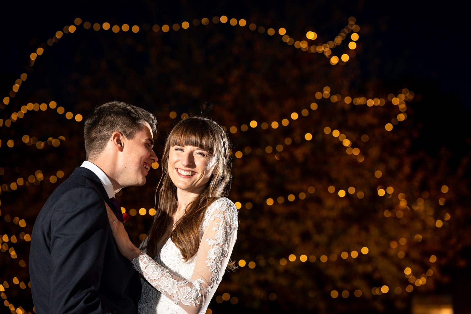 A smiling bride and groom sharing a joyful moment at their wedding with festive lights in the background.