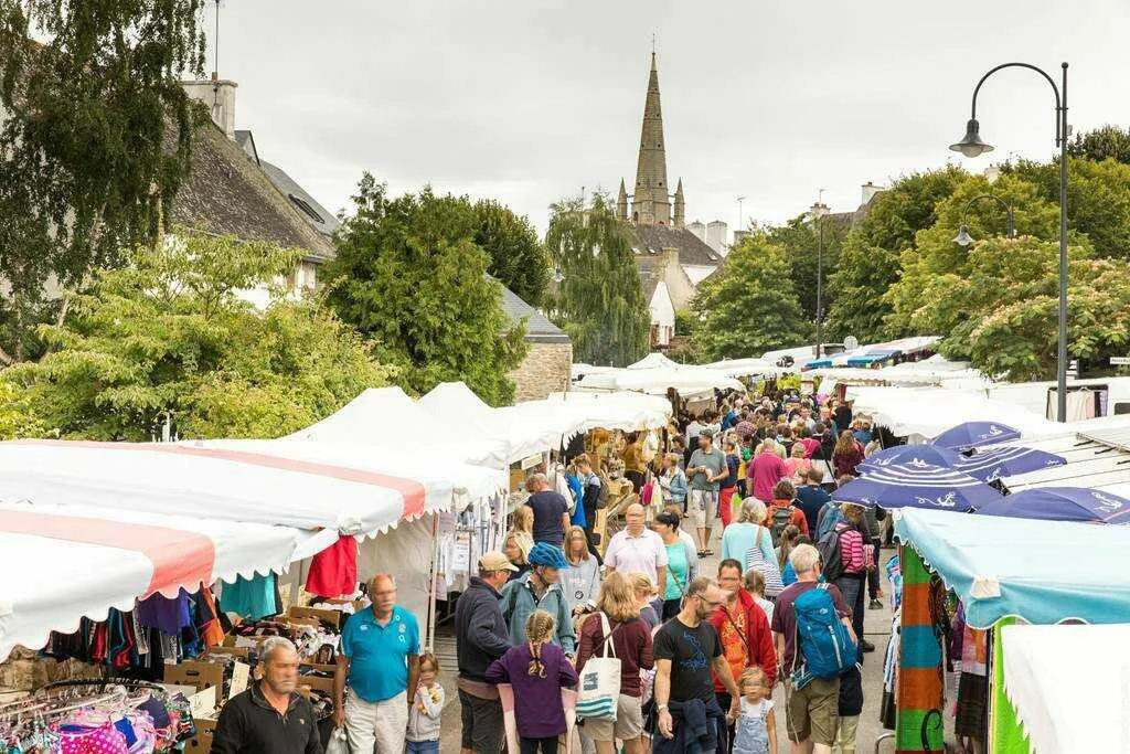 A crowded aisle at Carnac market, amongst the stalls, with the church tower in the background