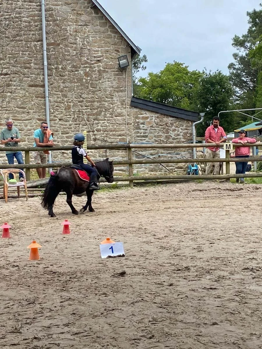A child riding a pony in the arena at the Centre Équestre des Menhirs, with parents watching from the sidelines