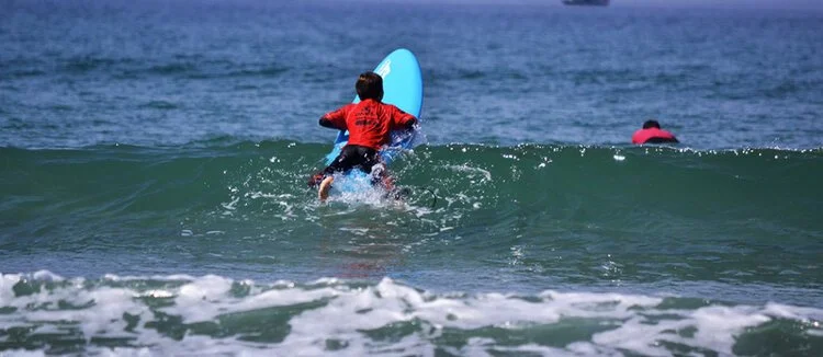 Kind im roten Lycra-Shirt paddelt auf einem blauen Surfbrett auf eine Welle zu, Surfkurs in der Bucht von Quiberon bei Carnac