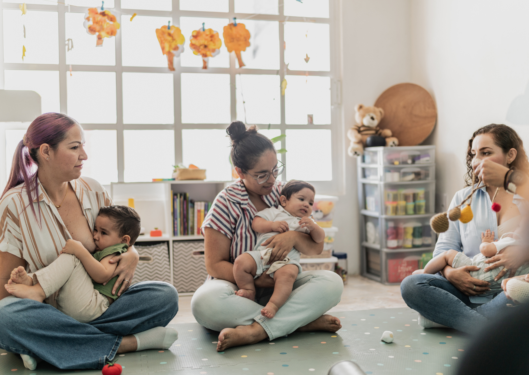 Three women sit cross-legged on the floor of a playroom with two babies, two women on the left and one on the right, engaging in conversation. The room has a large window with colorful paper trees hanging above, a bookshelf, a plastic storage drawer, and plush toys, including a teddy bear.