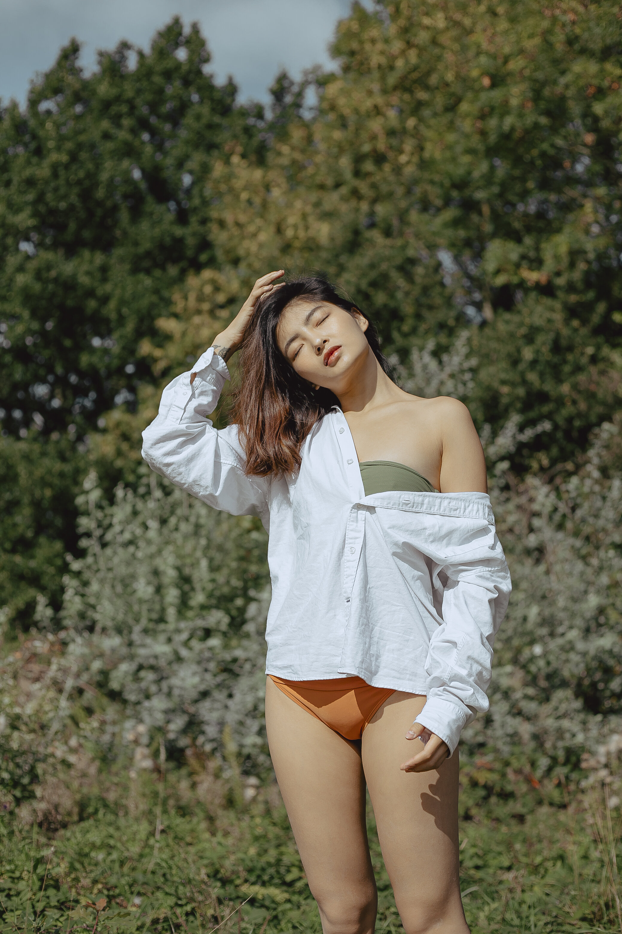 Young woman with long brown hair in outdoor setting, wearing an off-shoulder green top, a white oversized shirt, and orange bikini bottom, with eyes closed and one hand on her head, standing among trees and greenery.