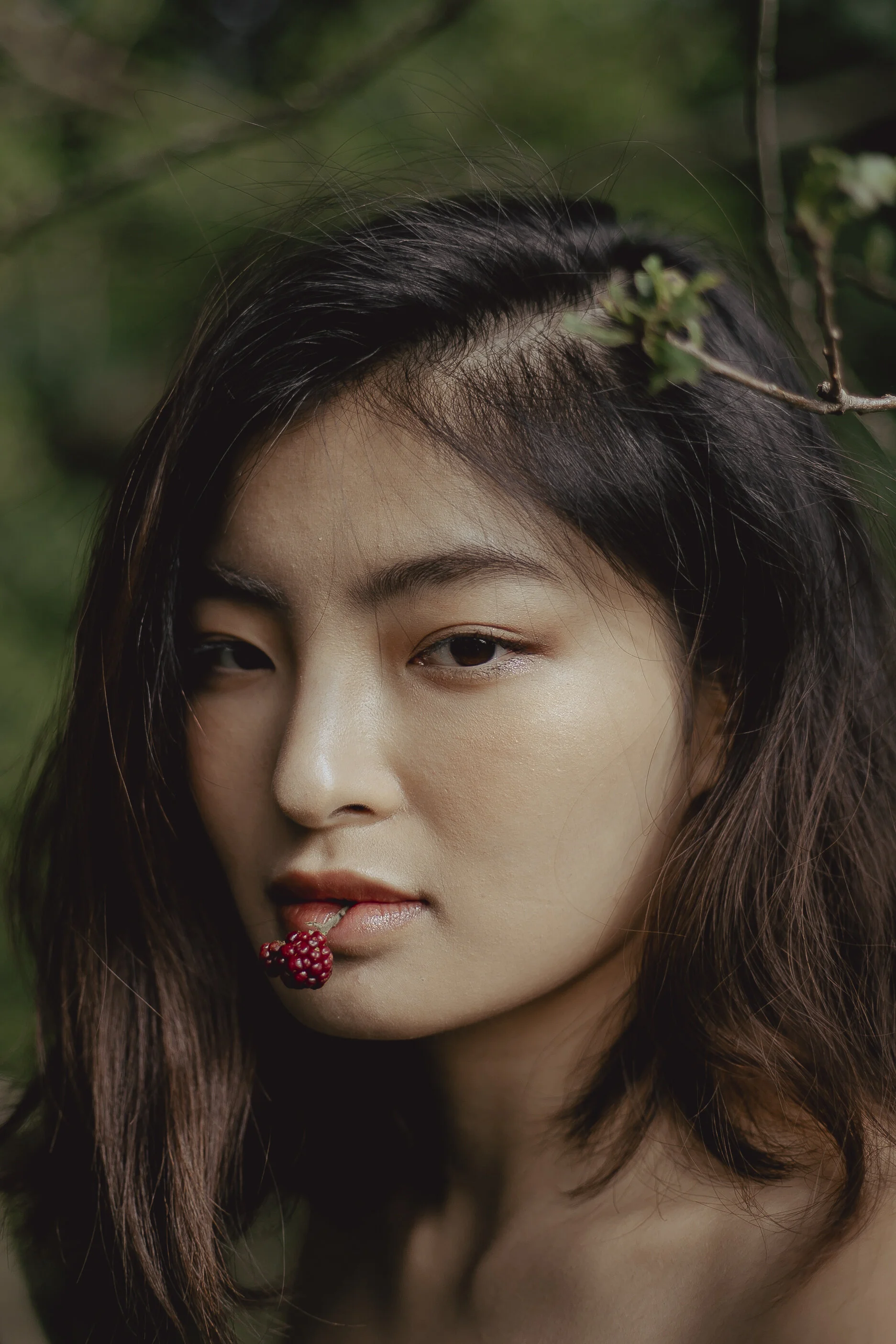 A young woman with dark hair, partially obscured face, and a blackberry in her mouth, surrounded by green foliage.