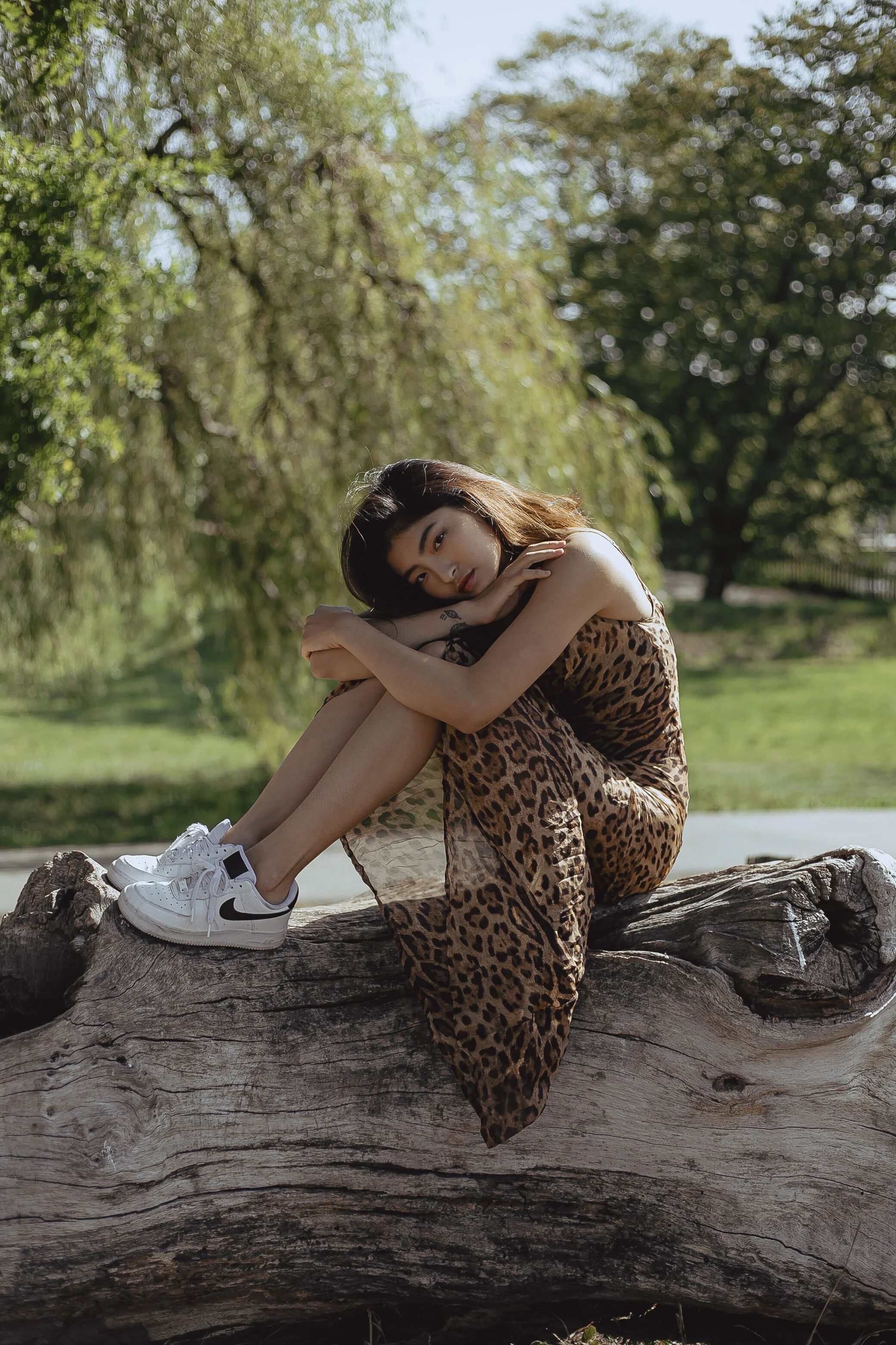 A young woman in a leopard print dress and Nike sneakers sitting on a large fallen tree log outdoors, with trees and sunlight in the background.