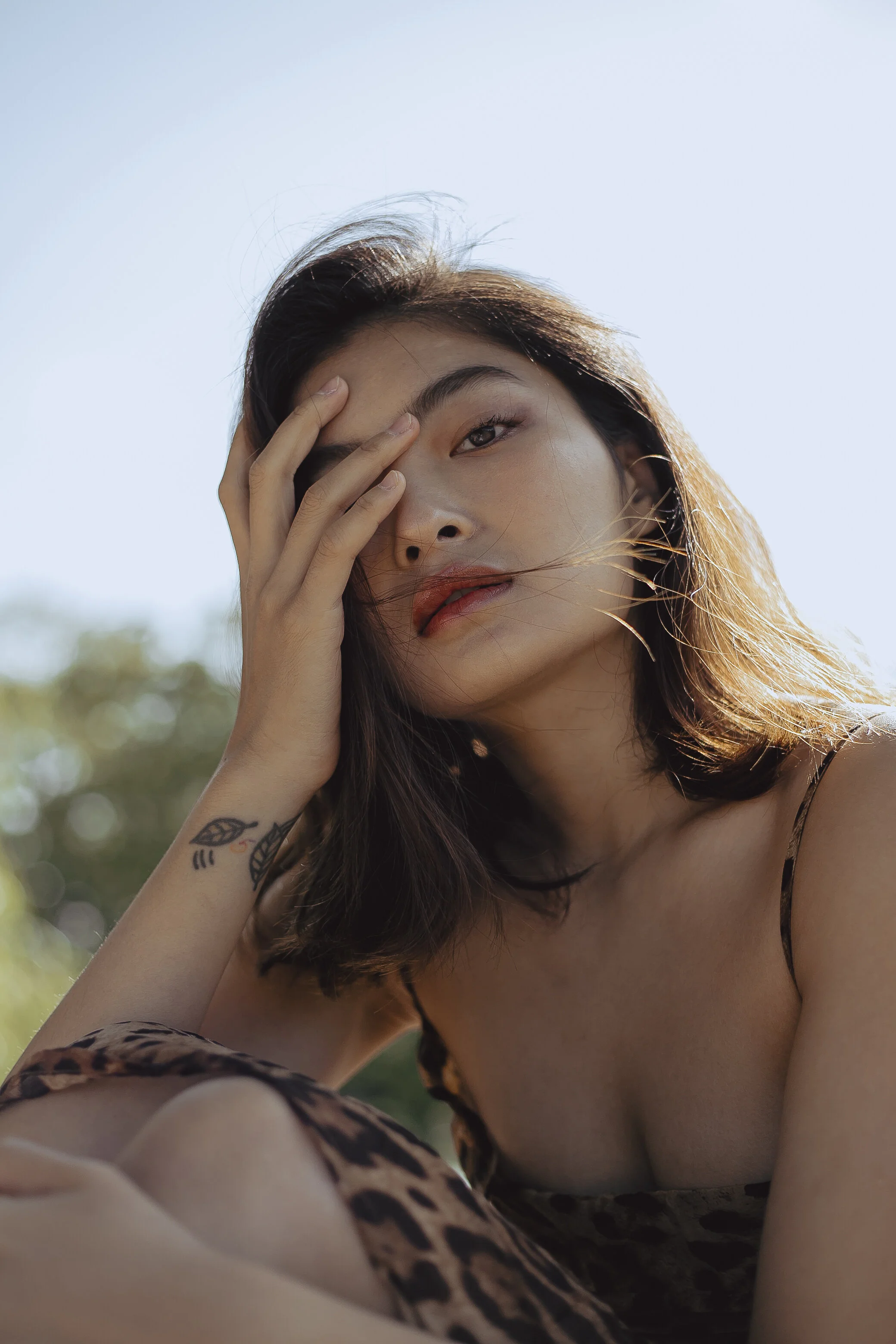 Close-up of a woman with shoulder-length dark hair, wearing a leopard-print top, holding her face with her left hand, outdoors with a blue sky and blurred greenery in the background.