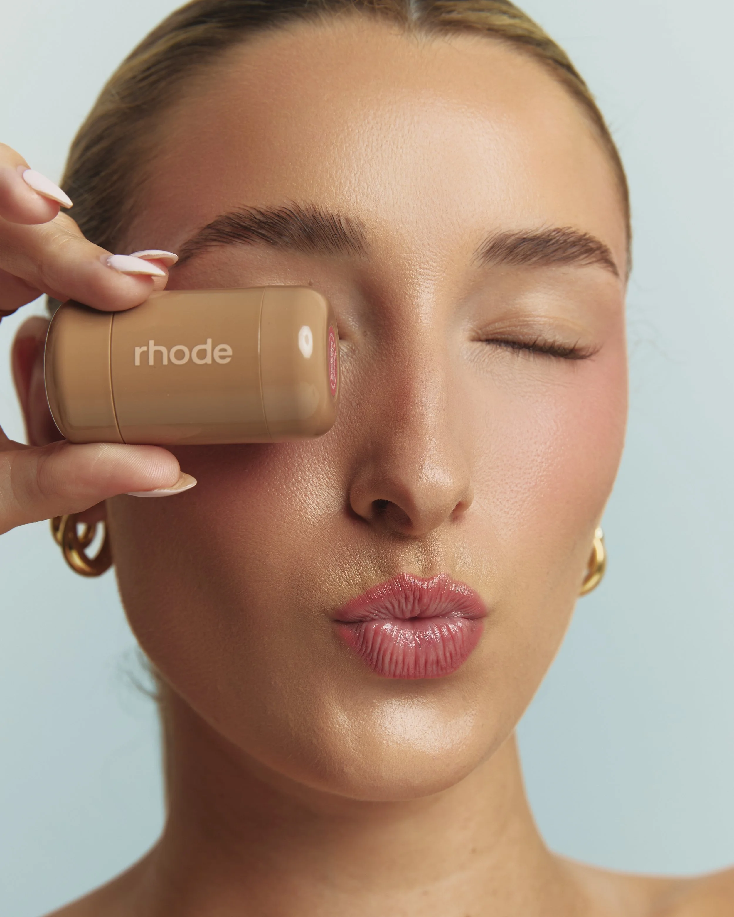 A woman with light brown hair and gold hoop earrings is holding a beige makeup sponge labeled "rhod" against her right temple. She has minimal makeup, neatly groomed eyebrows, and her eyes are closed.