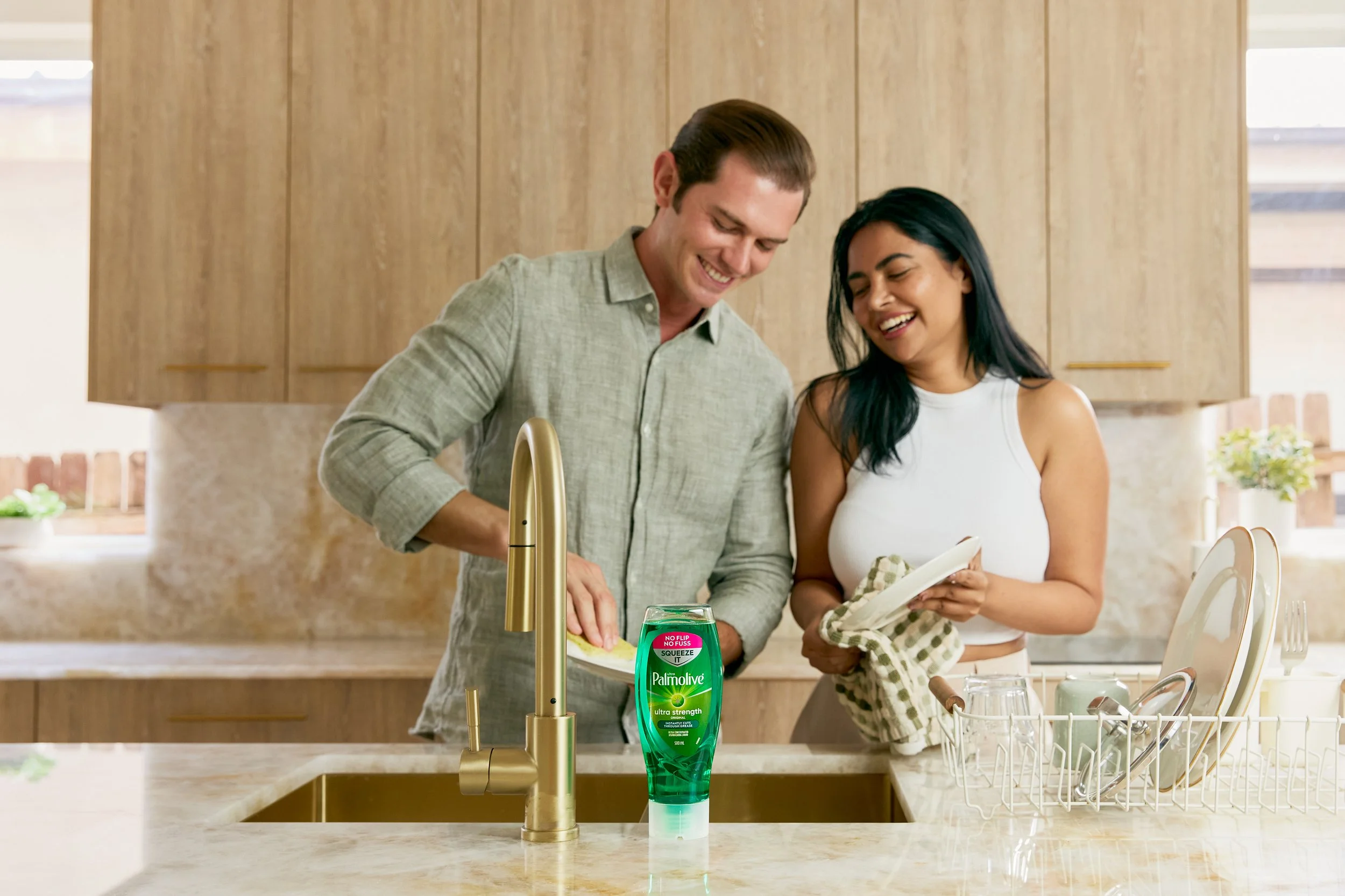 A man and woman standing at a kitchen sink, smiling and washing dishes together. The woman is holding a dish and a dish towel, the man is at the sink, and there is a bottle of Palmolive dish soap on the counter.