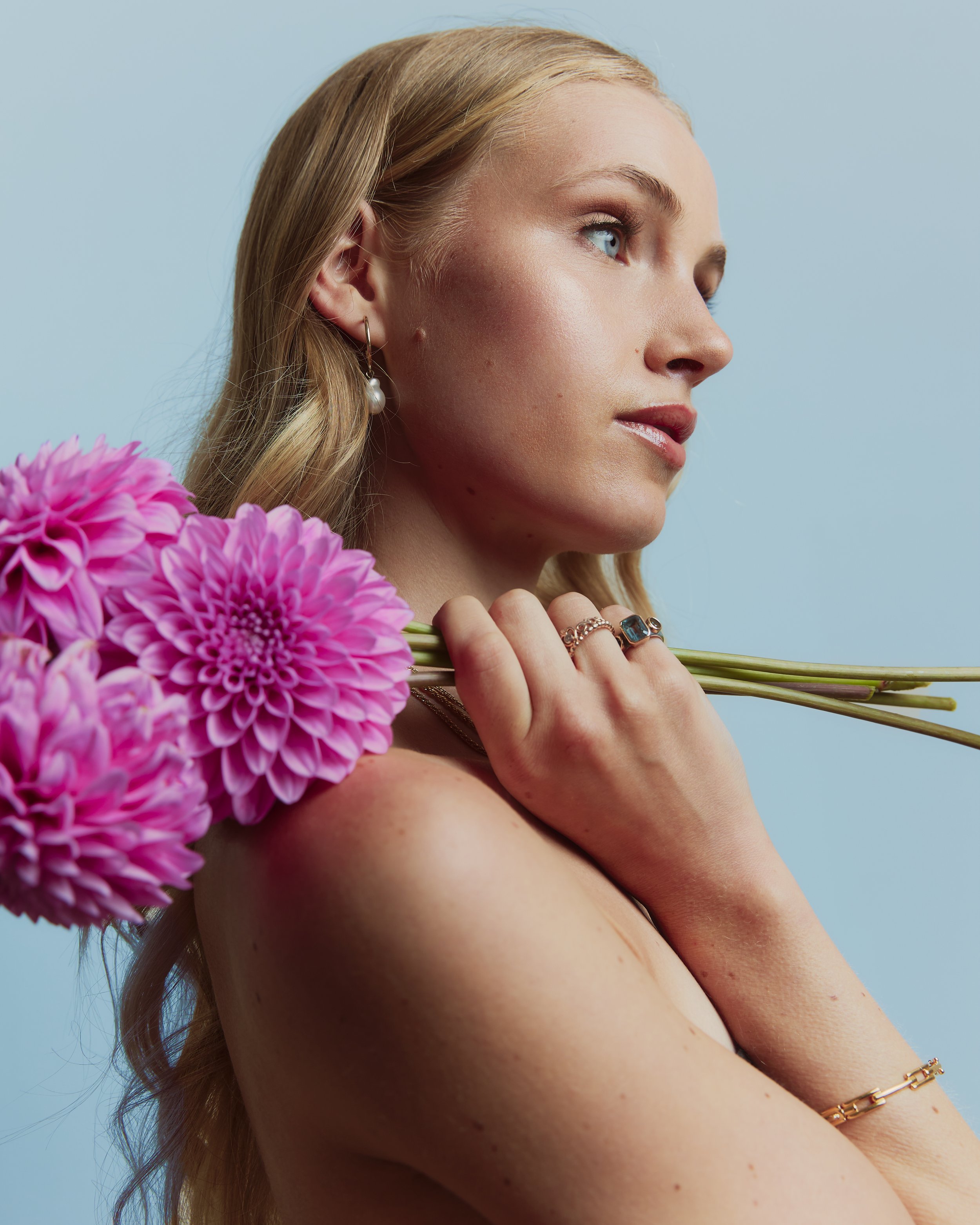 A woman with long, wavy blonde hair and blue eyes looking to the side, holding pink flowers, wearing pearl earrings and gold rings, against a light blue background.