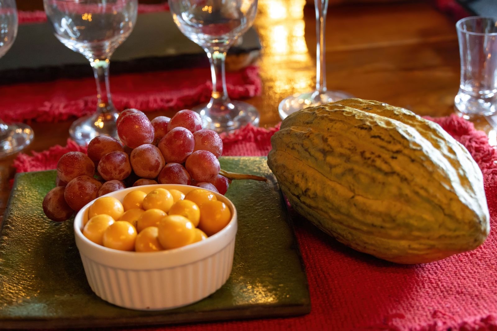 A table setting with grapes, yellow cherry tomatoes in a small bowl, butternut squash, empty wine glasses, and red placemats, on a wooden table.