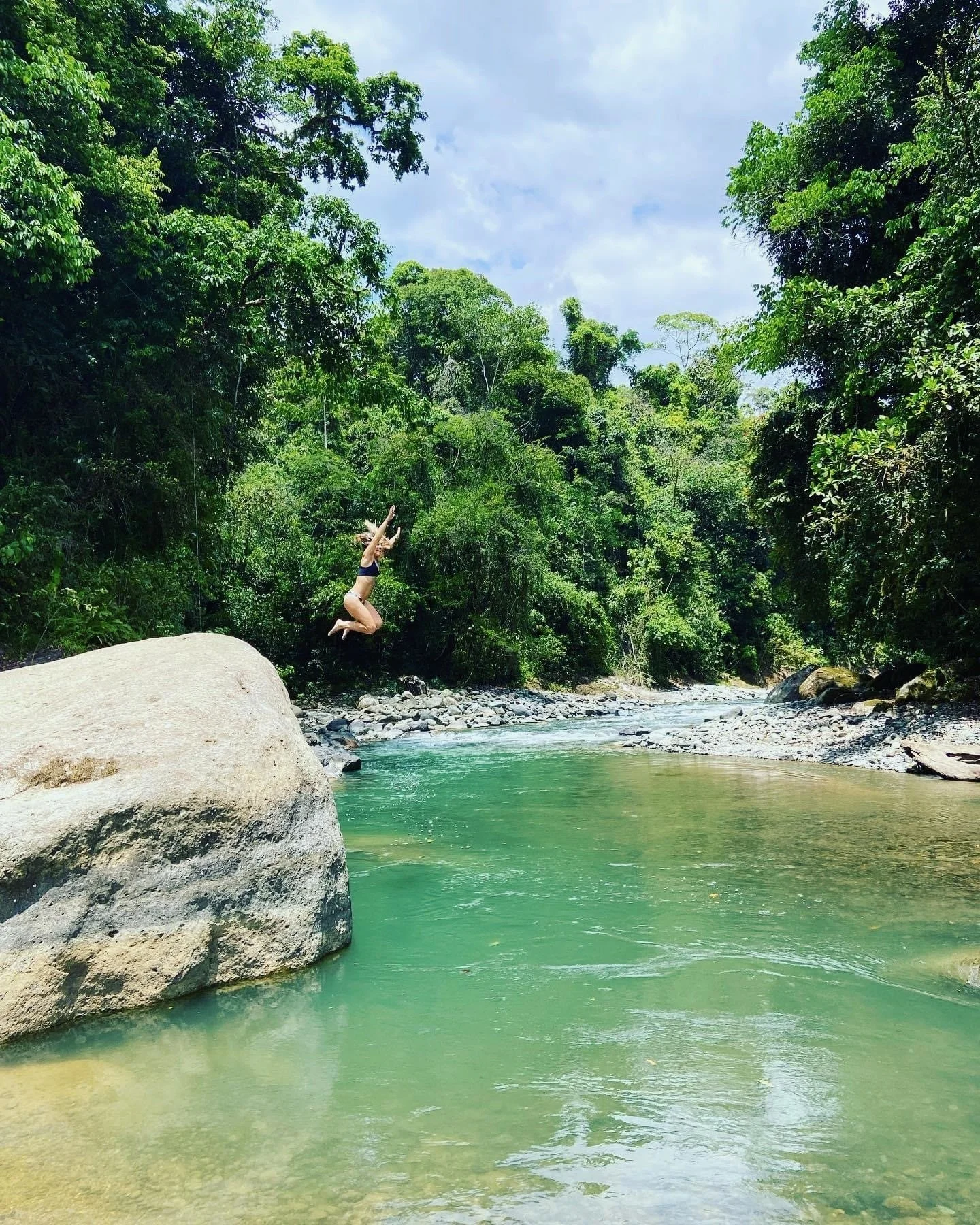A person jumping into a river surrounded by lush green trees with a cloudy sky overhead.