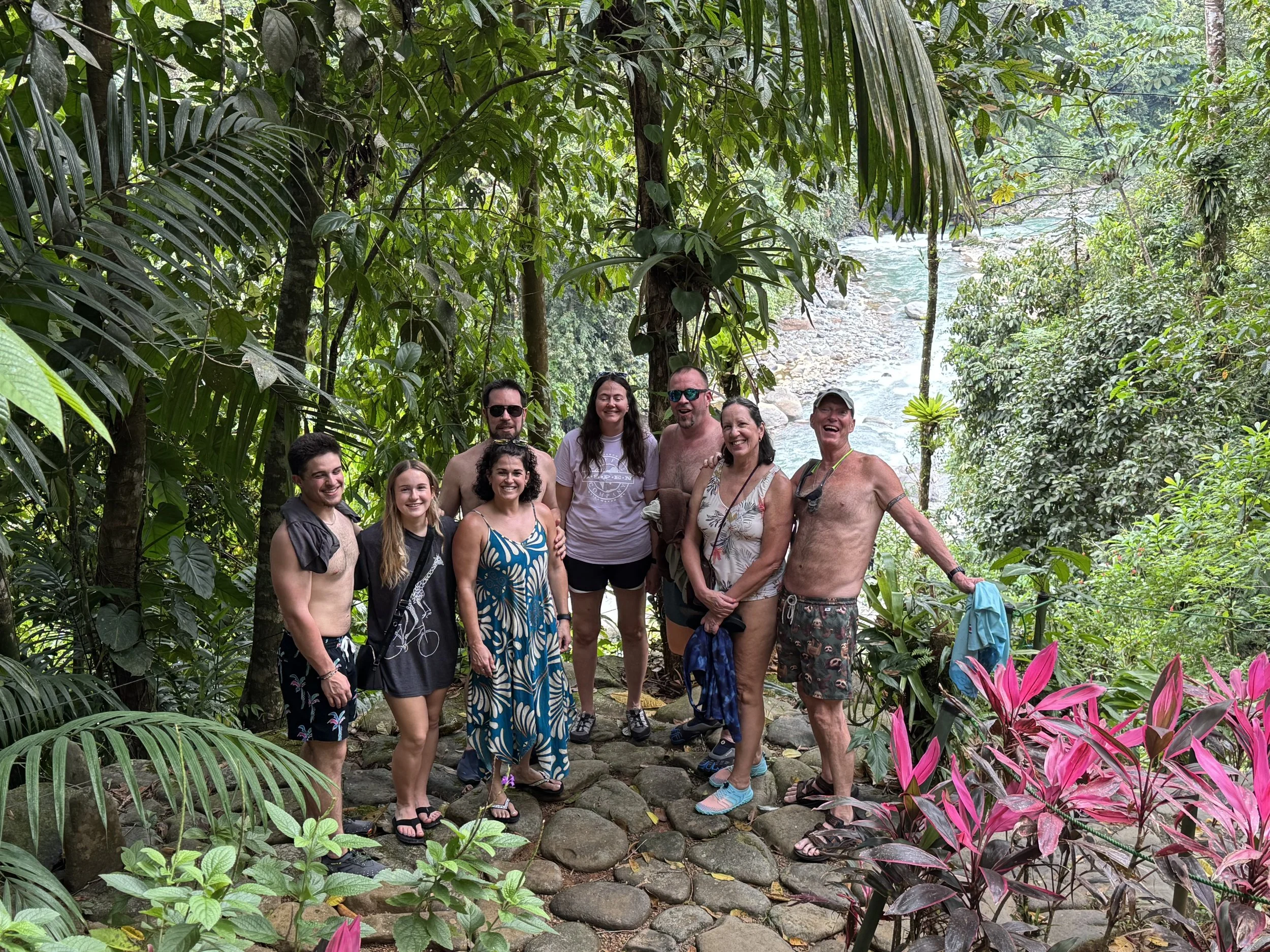 A diverse group of ten people standing together in a lush tropical jungle near a river, smiling and enjoying their outdoor adventure.