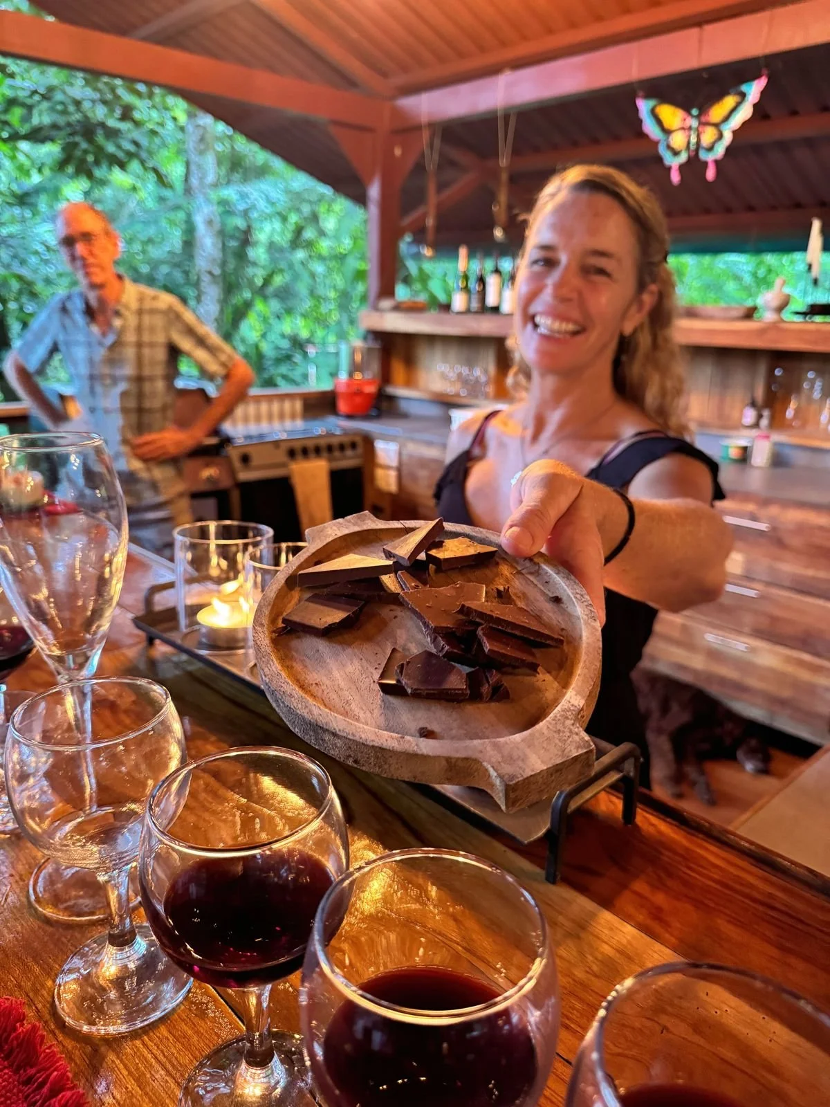 A happy woman extending a wooden plate of chocolate pieces towards the camera, with glasses of red wine and a man in the background in a rustic, outdoor kitchen setting.