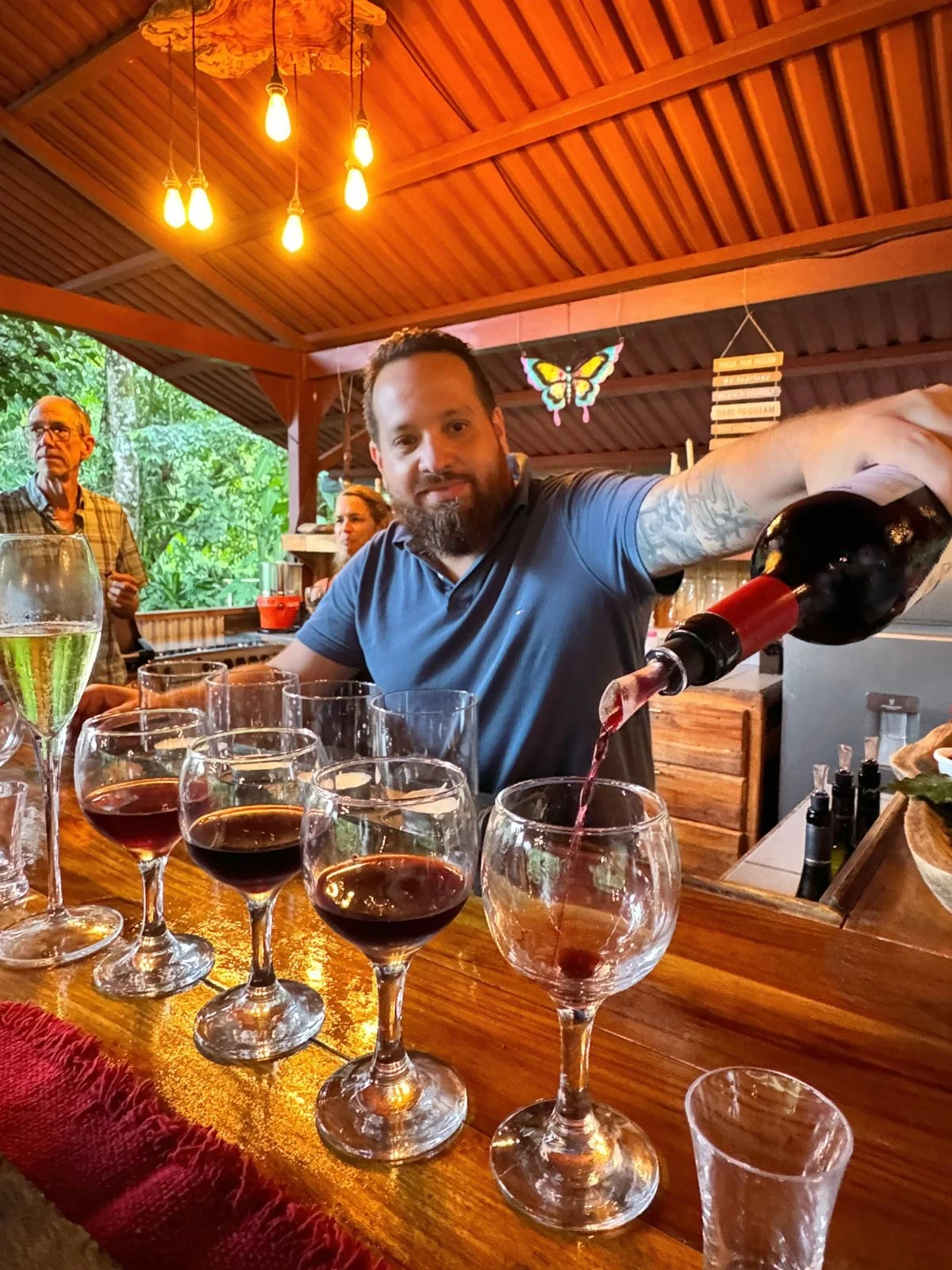 A man with a beard in a blue shirt pours red wine into a glass at a wooden bar with other glasses of wine, while a background of wood-paneled ceiling and windows showing greenery is visible.