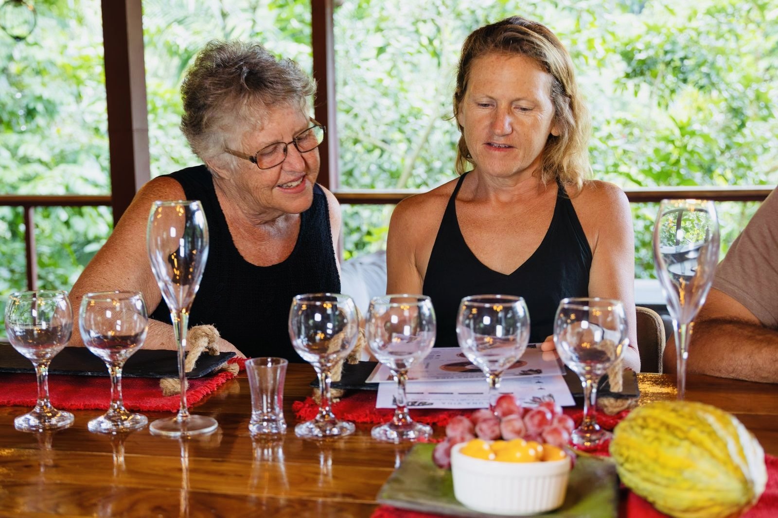 Two women sitting at a table with empty glasses, looking at a menu or book, in a bright outdoor setting with green foliage in the background.