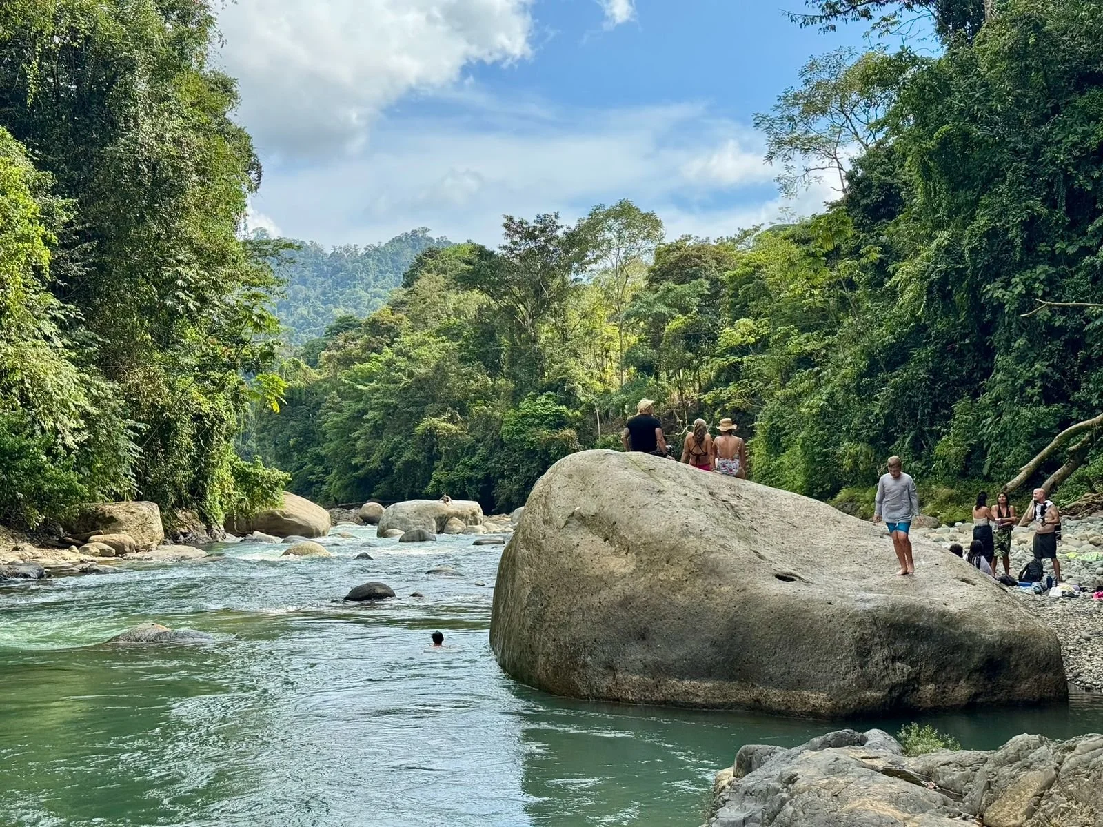 People relaxing and sitting on rocks by a river in a lush green forest with mountains in the background.