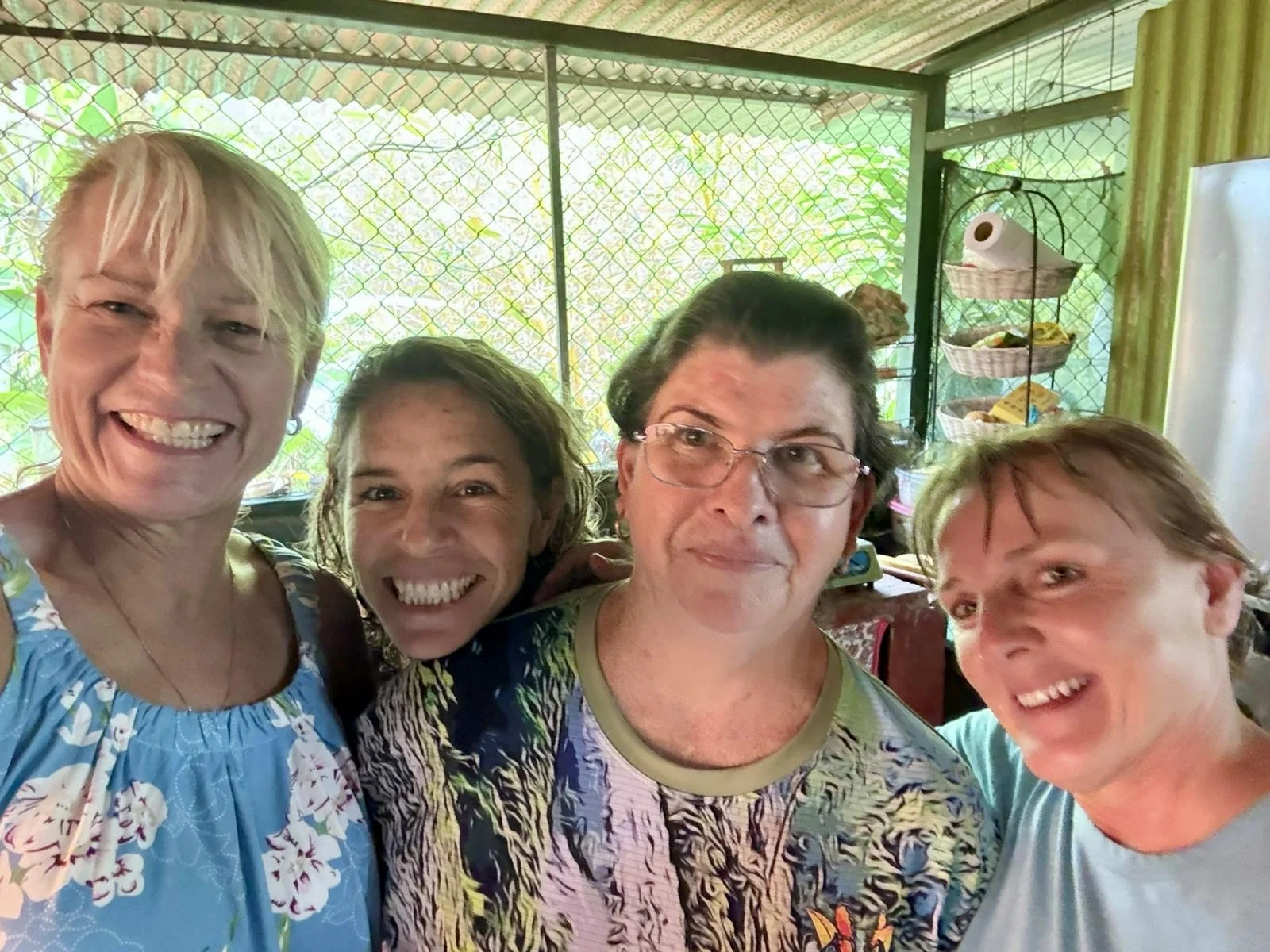 Four smiling women in a close group selfie, standing inside a room with a screened window in the background.