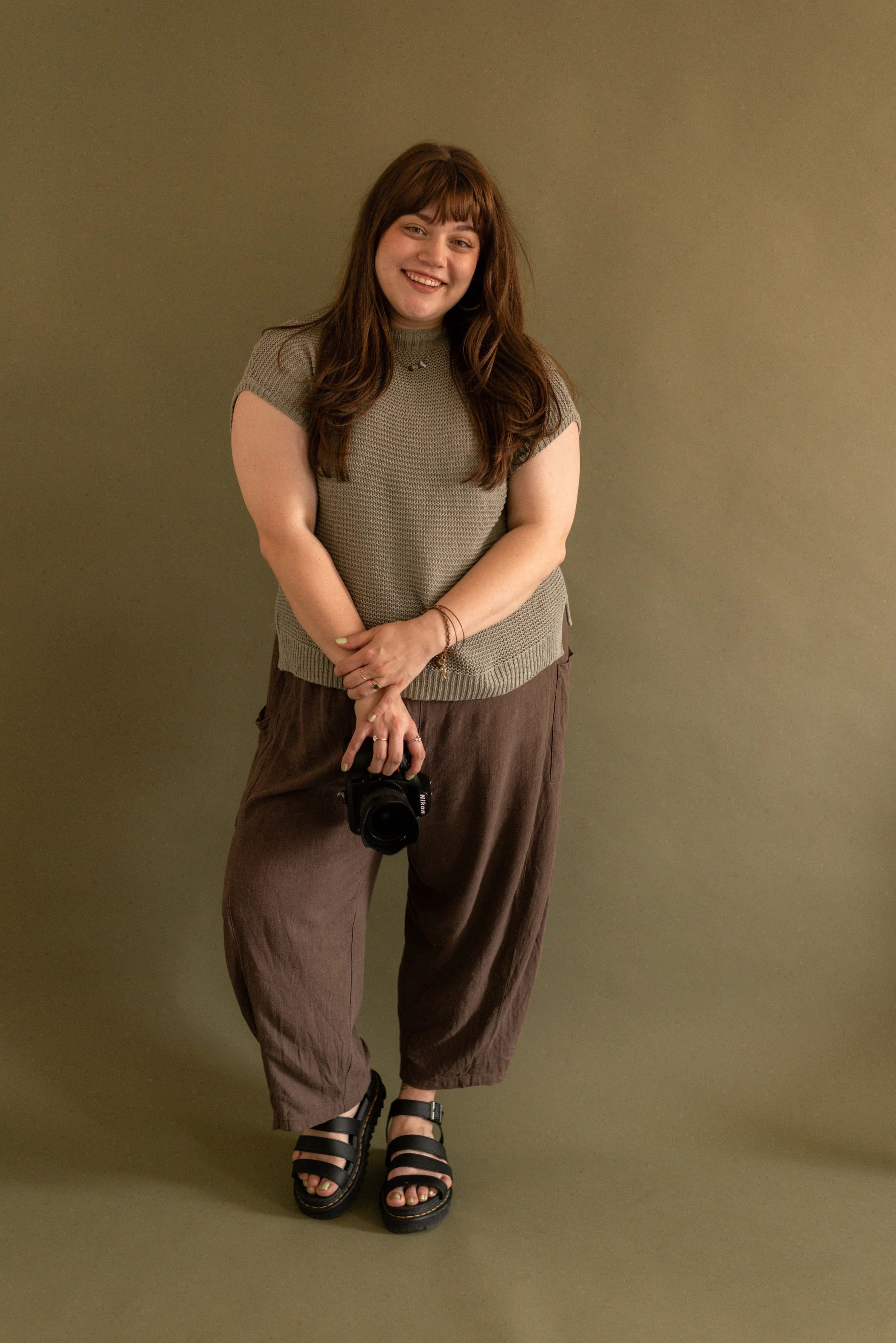 an indiana wedding and boudoir photographer stands posing in her natural light photography studio in front of an olive green backdrop. She has her hands clasped on her camera and is smiling directly into the camera. Her face is warm and inviting.