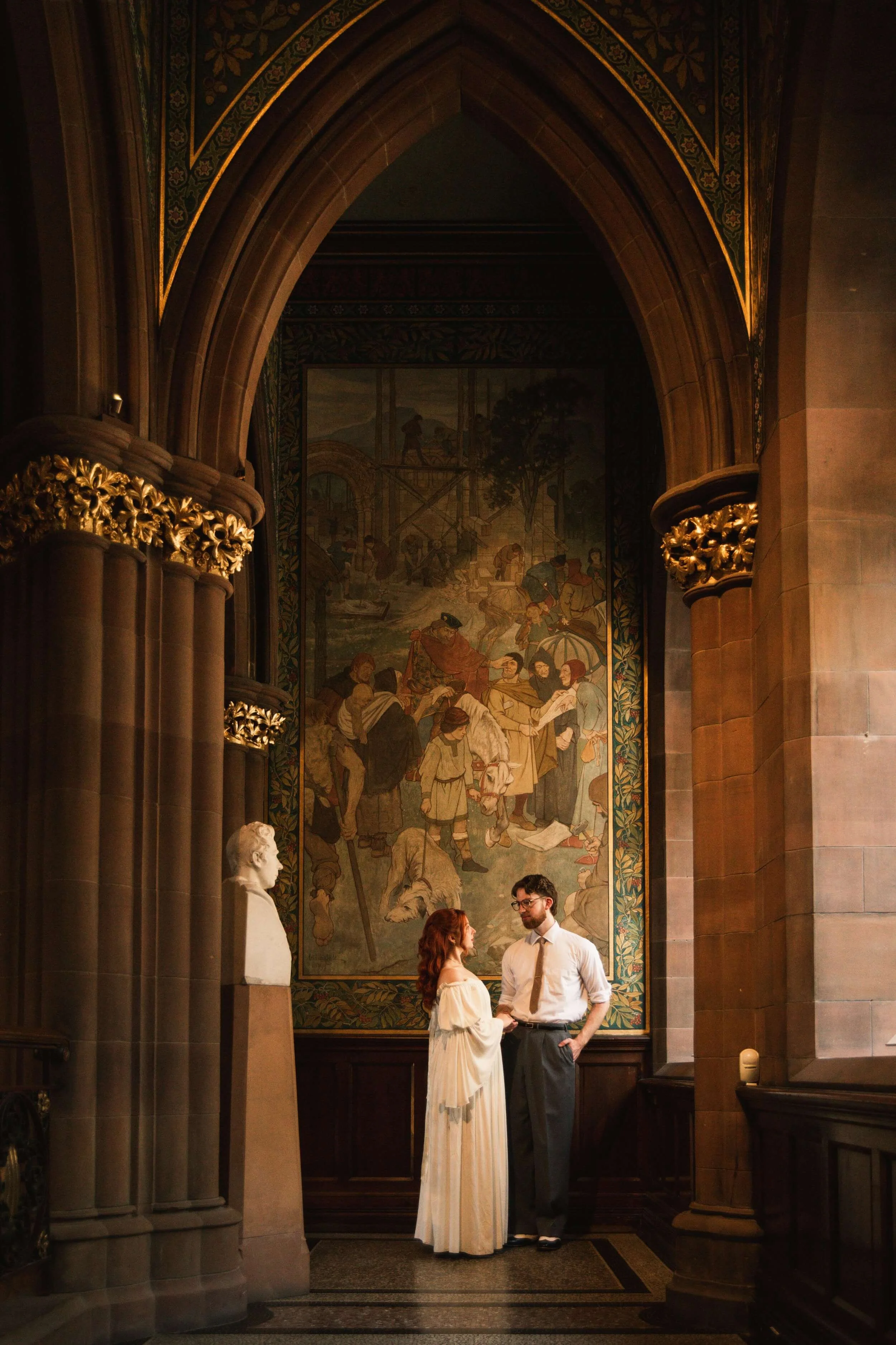 Abride in a white dress and her groom in a white button up shirt and tie stand together under a large archway in the National Portrait Gallery of Scotland, with a fresco mural and a white bust behind them.