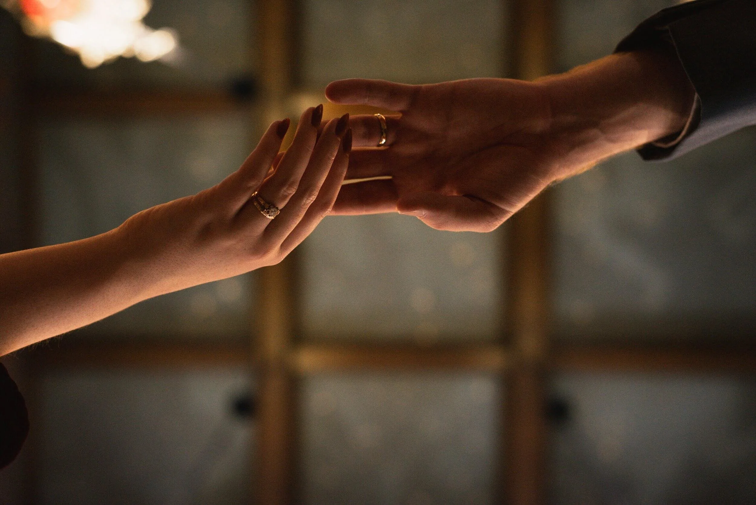 Two hands reaching toward each other in a gentle gesture, both wearing new wedding rings, with a softly lit background and warm tones of the ceiling above.